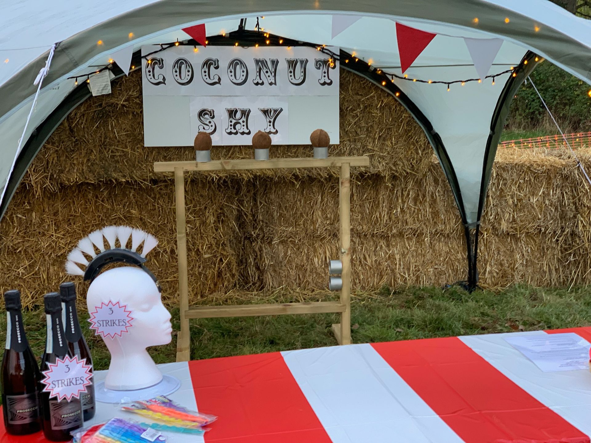 Coconut shy game at a fair. Coconut stand in front of hay bales. Prizes on a red & white striped table.