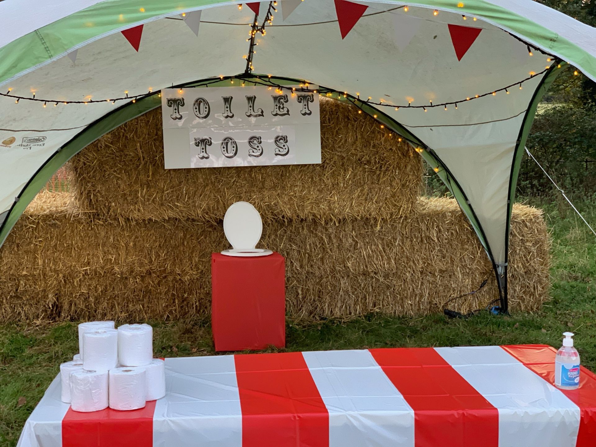 Carnival game booth with a toilet toss, hay bales, and red & white table.
