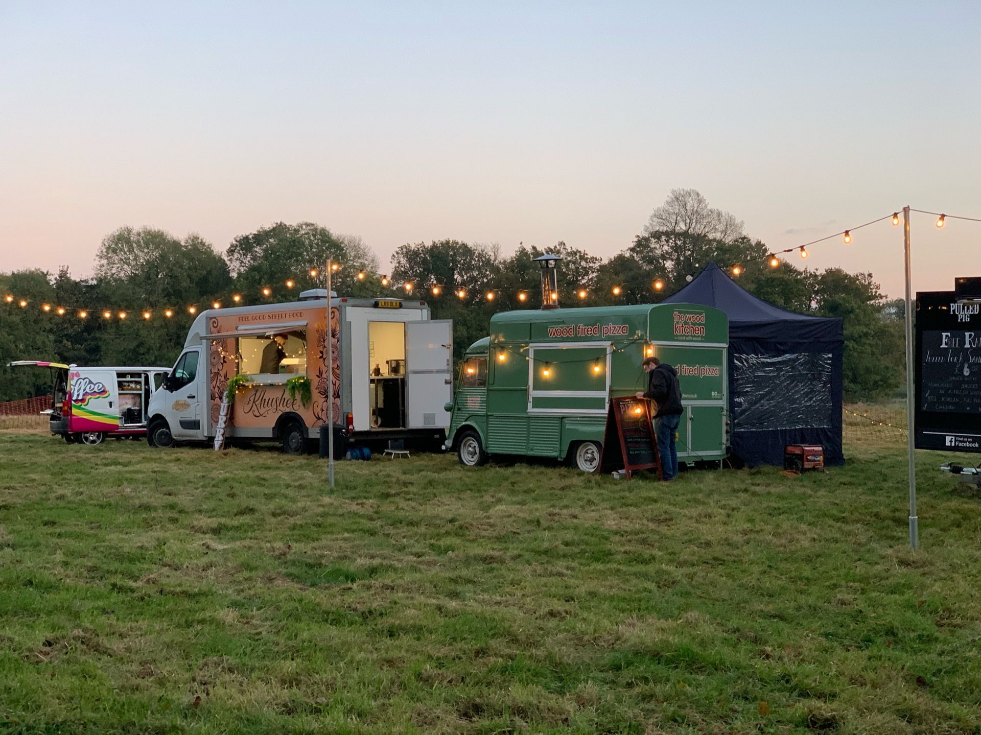 Food trucks set up in a grassy field, lit by string lights. Two trucks are open for business.