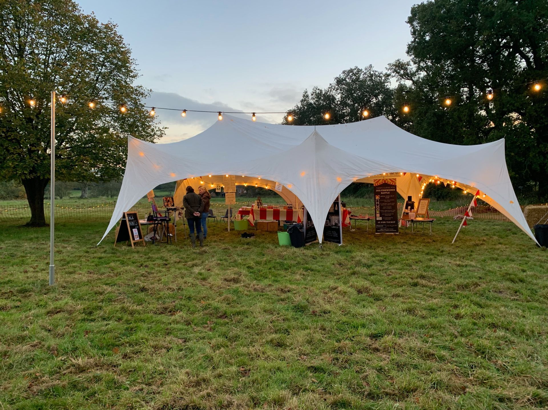 White tent set up in a grassy field, strung with lights, people milling about.