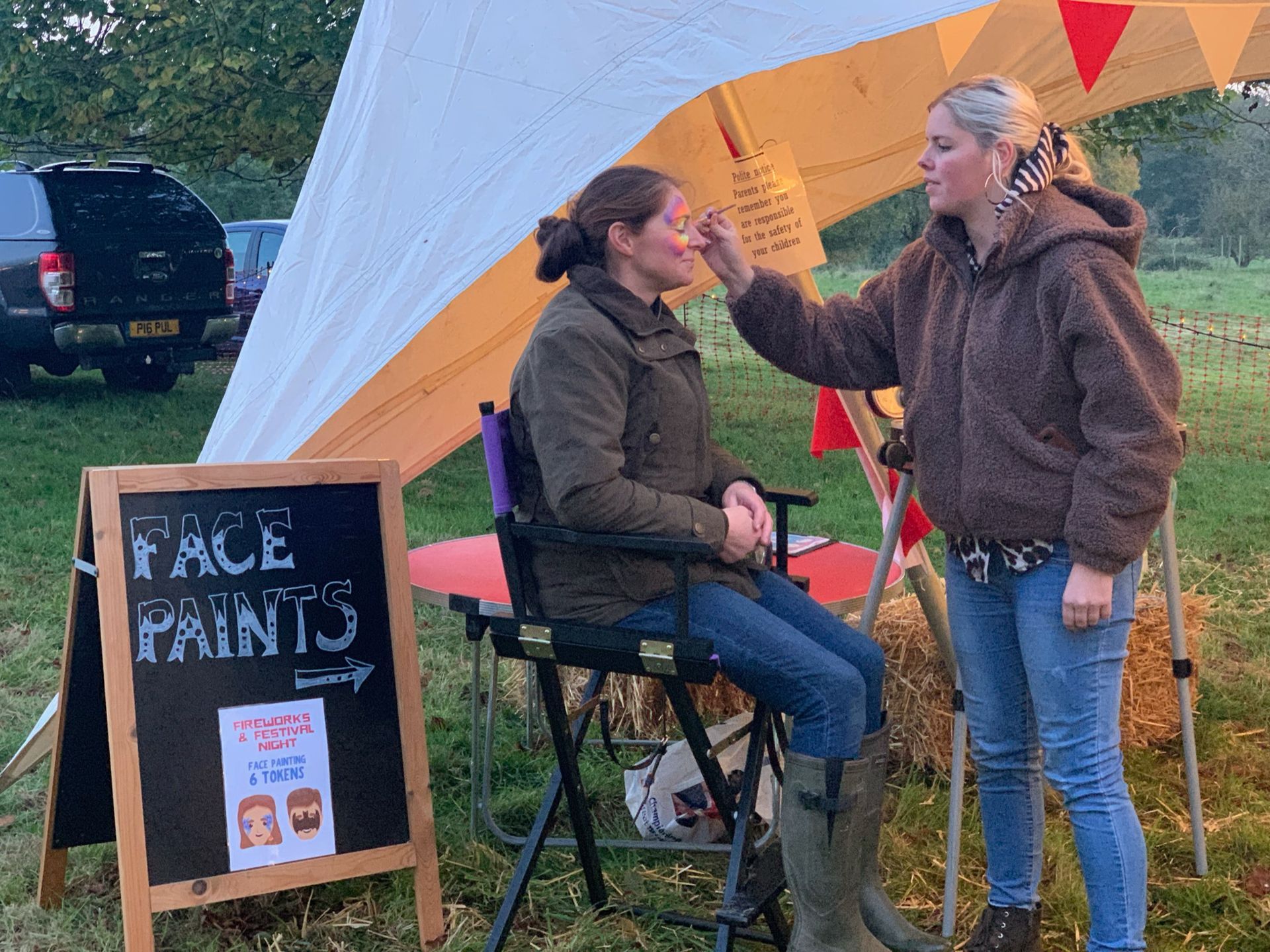 Woman getting face painted at an outdoor event. Tent, sign, and parked vehicle visible.