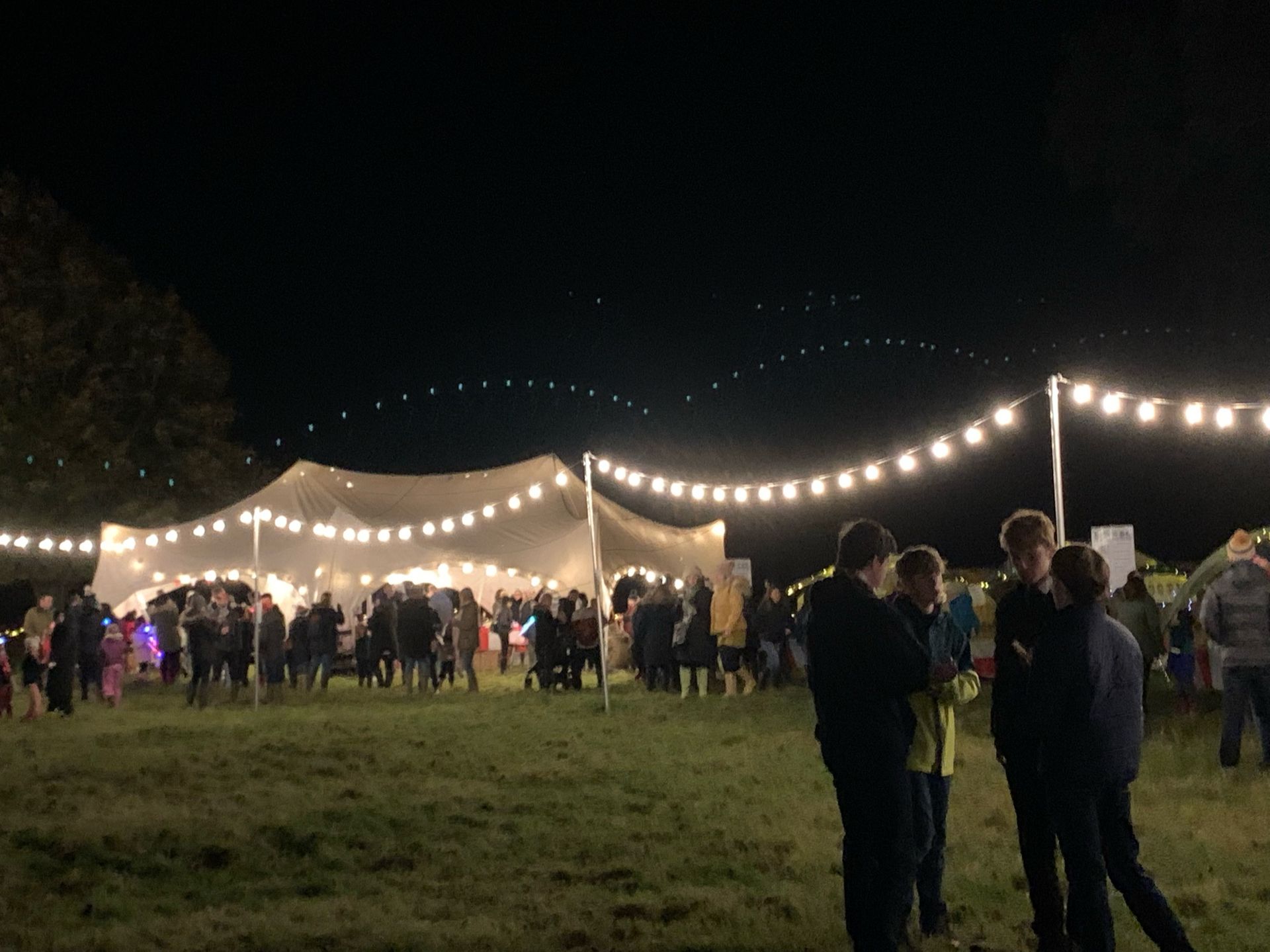 Nighttime outdoor event with a tent, string lights, and a crowd of people.