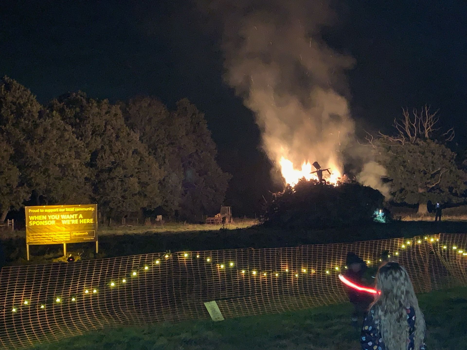 Large bonfire burning at night in a field, with people watching and string lights.