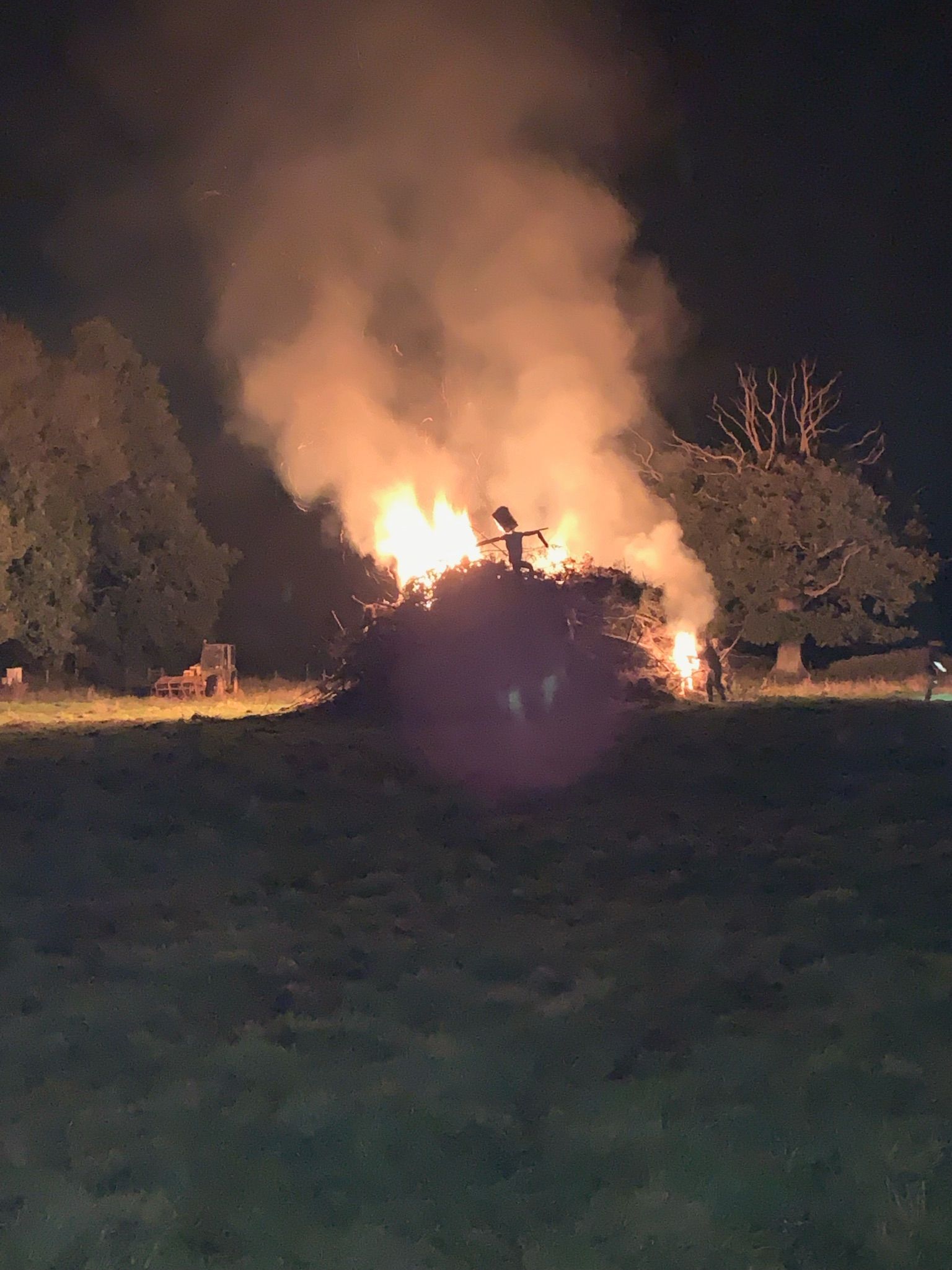 Large bonfire burning in a field at night; flames and smoke visible, trees in the background.