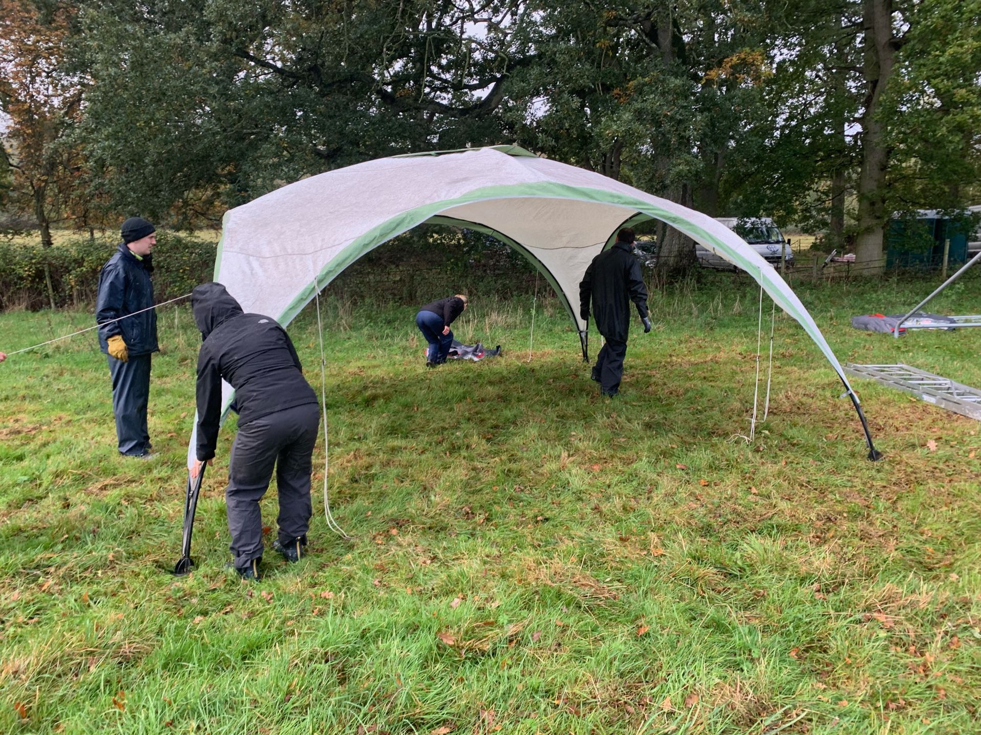 Four people setting up a white and green canopy in a grassy field near trees.