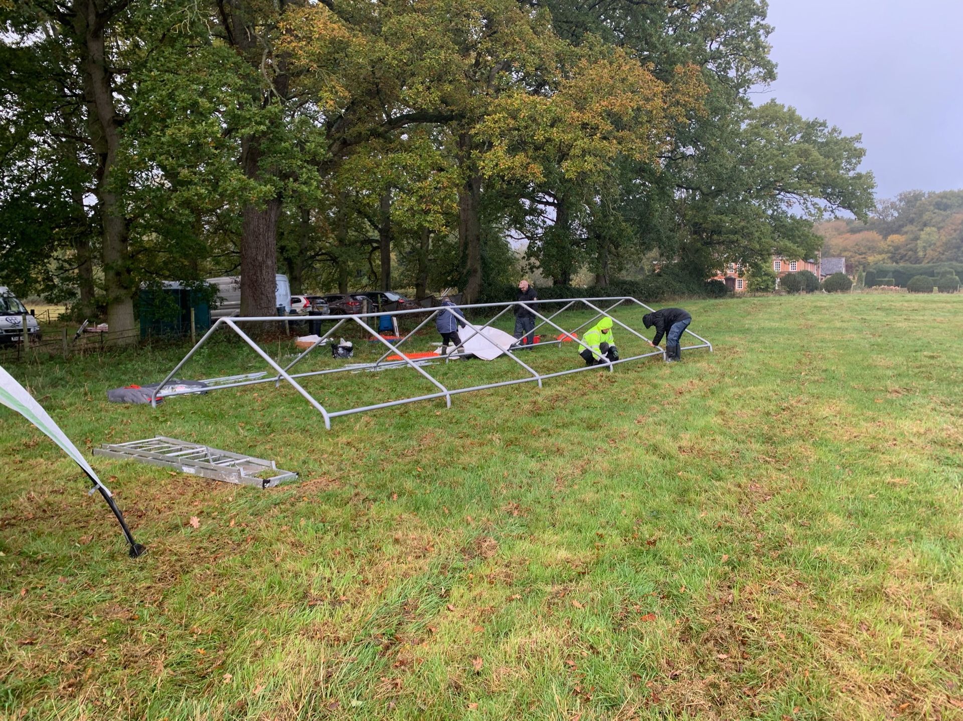 People assembling a silver framed tent on a grassy field, surrounded by trees under an overcast sky.