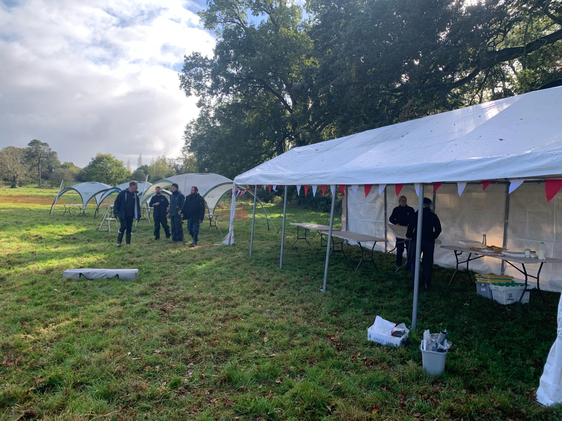 People gather at a field with tents and tables, preparing for an event, under a cloudy sky.