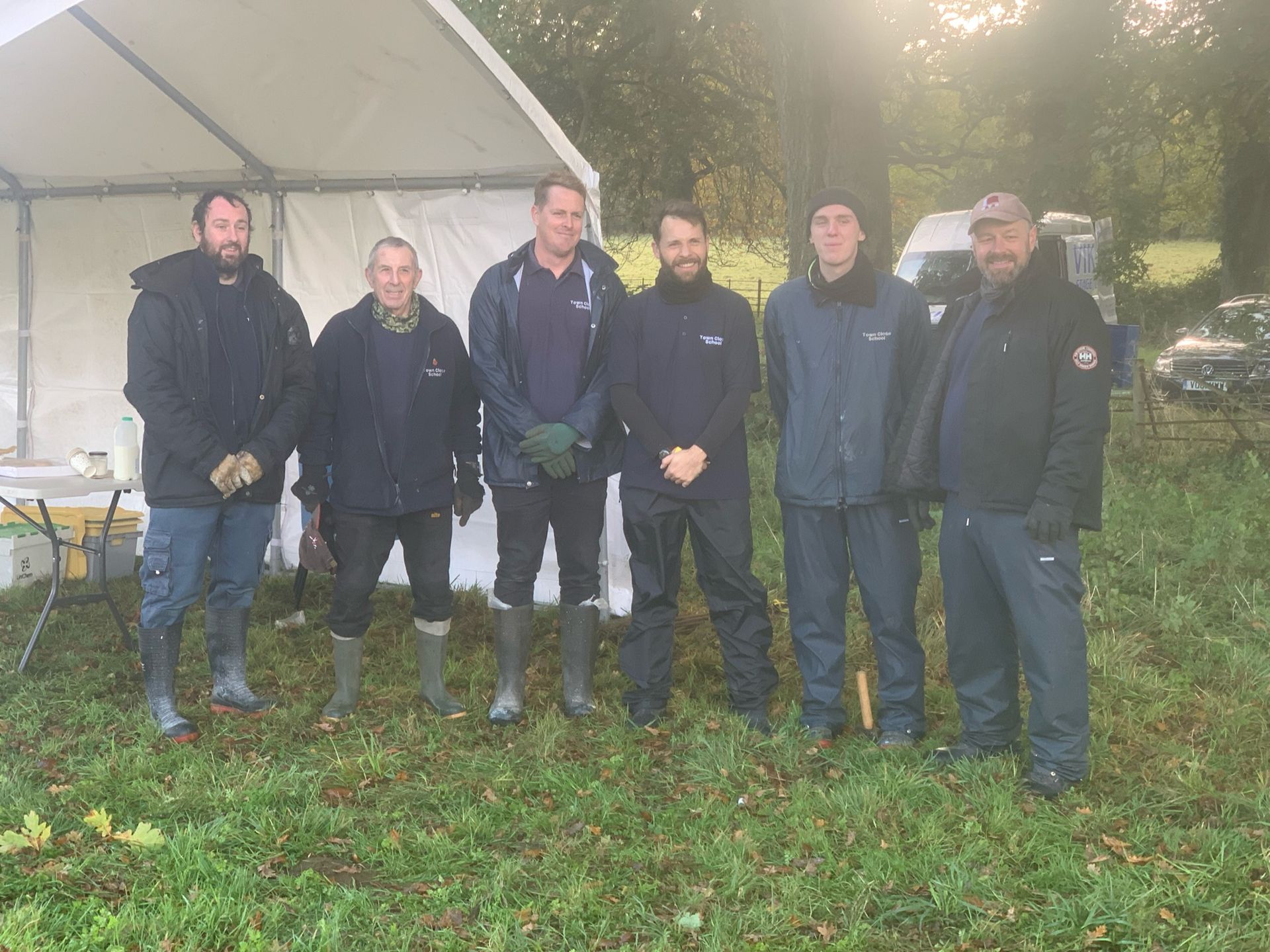 Six men stand in front of a white tent on a grassy field. Overcast, all wear outerwear.