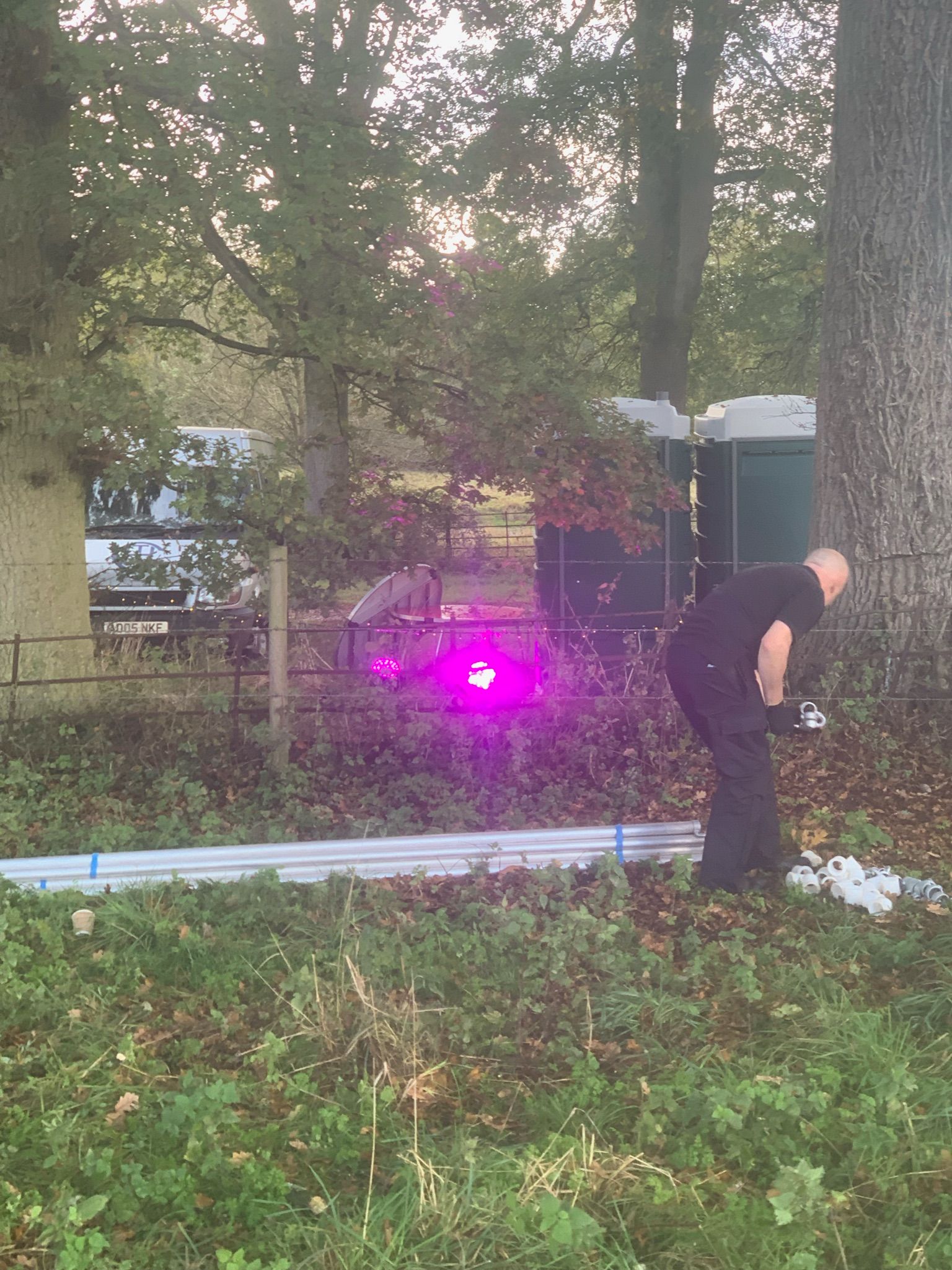Person in black bent over, near pink laser beam. Outdoor setting with trees, portable toilets.