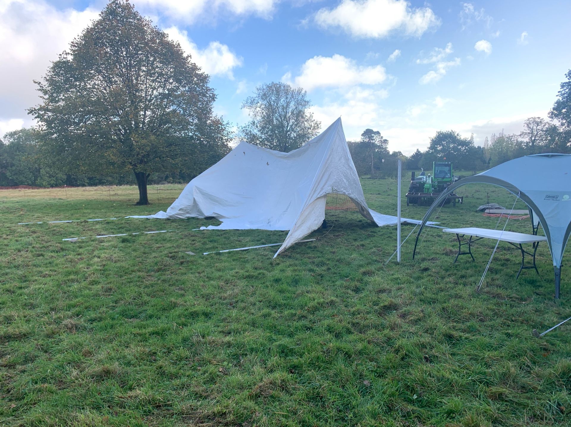 A white tent collapsed on a grassy field, with a gazebo and trees in the background.