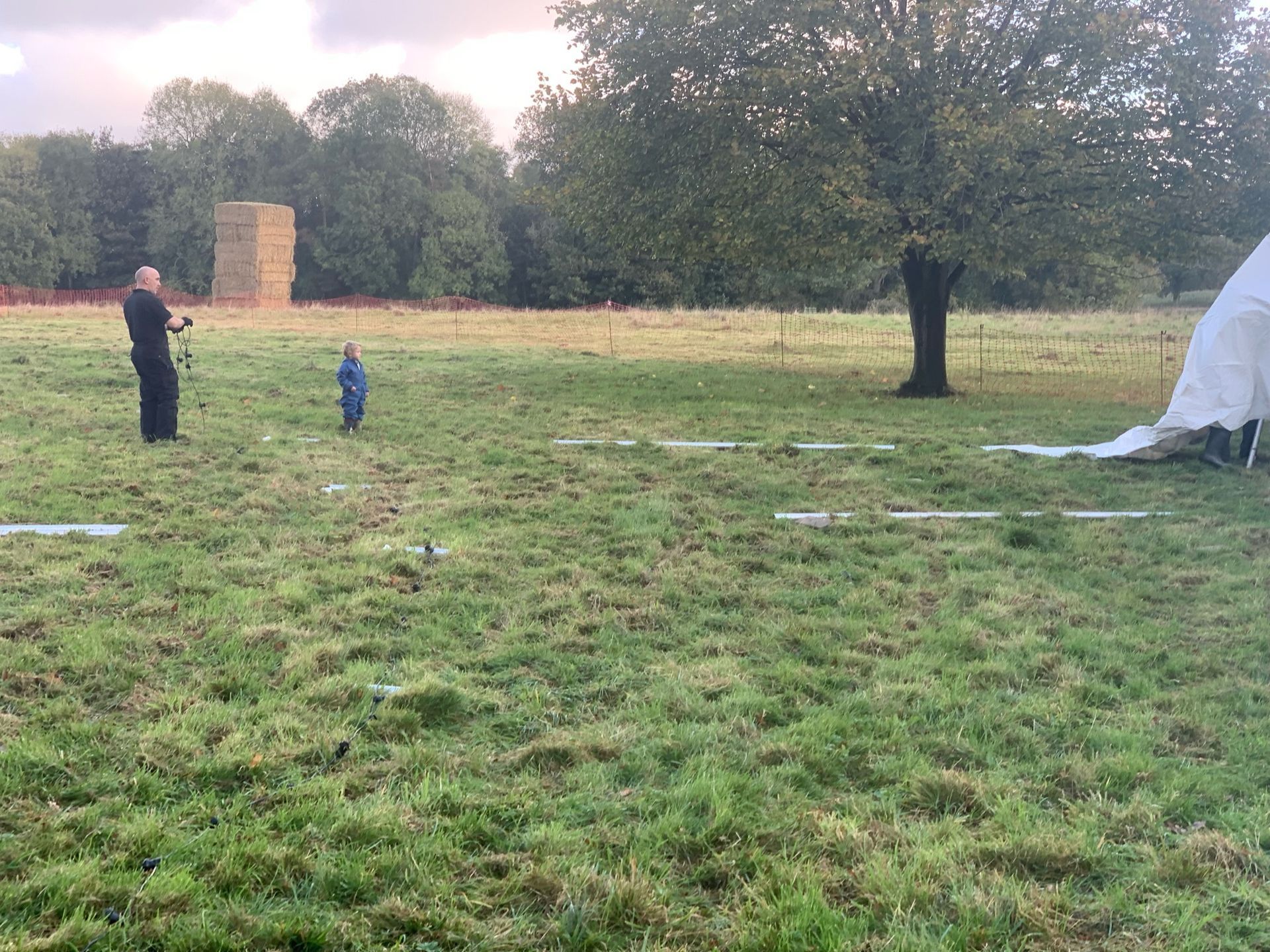 Man holding camera and small child in blue suit, in a grassy field. Tree and hay bales in the background.