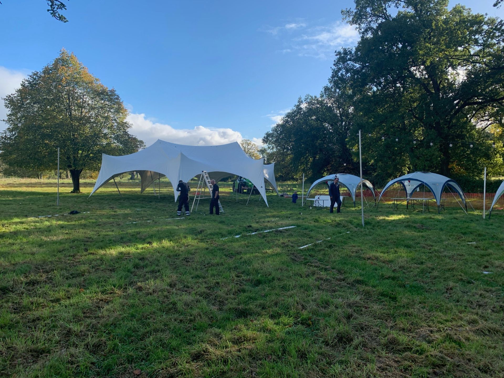 People setting up white tents in a grassy field, under a blue sky with trees.