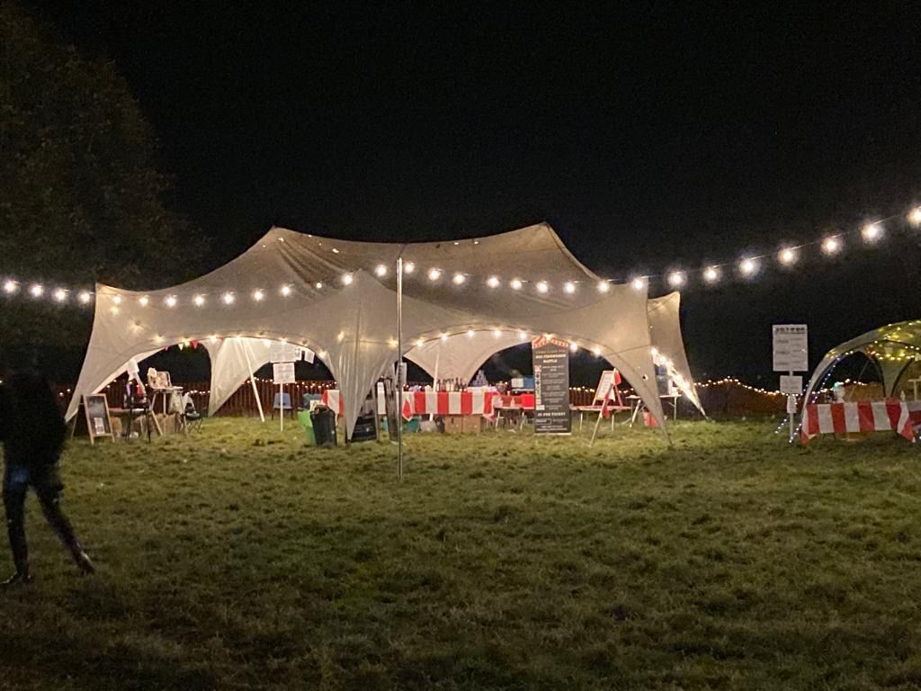 Night scene: Illuminated tent with string lights over tables on a grassy field, people present.