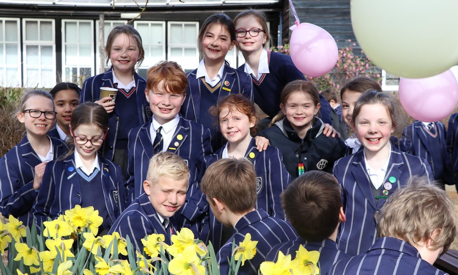 Group of school children in uniforms, smiling, near daffodils and balloons.