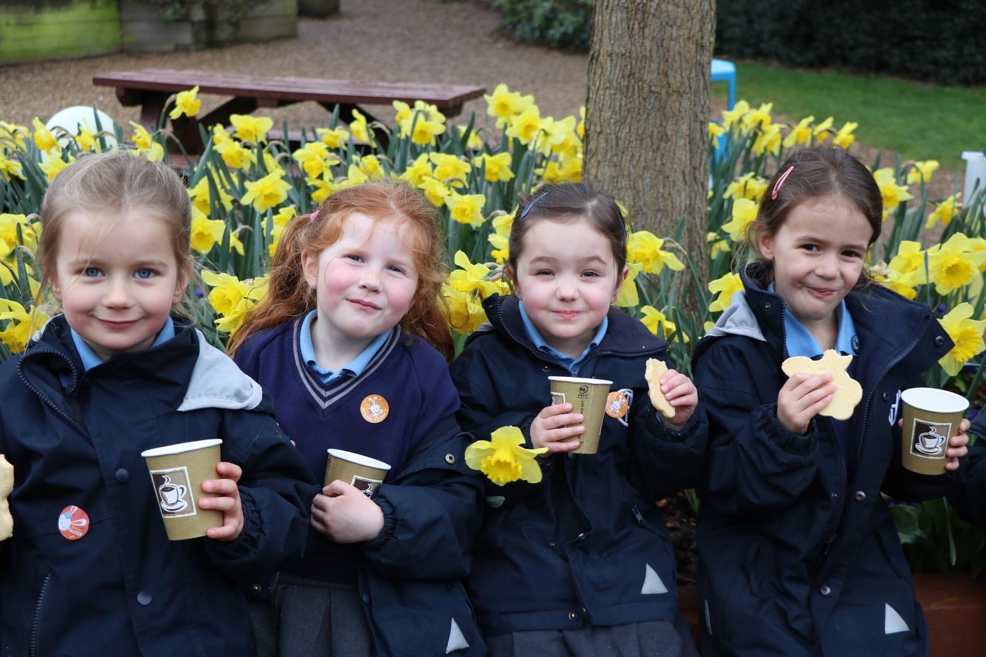 Four young girls in school uniforms enjoy drinks and cookies outdoors, smiling in front of yellow daffodils.
