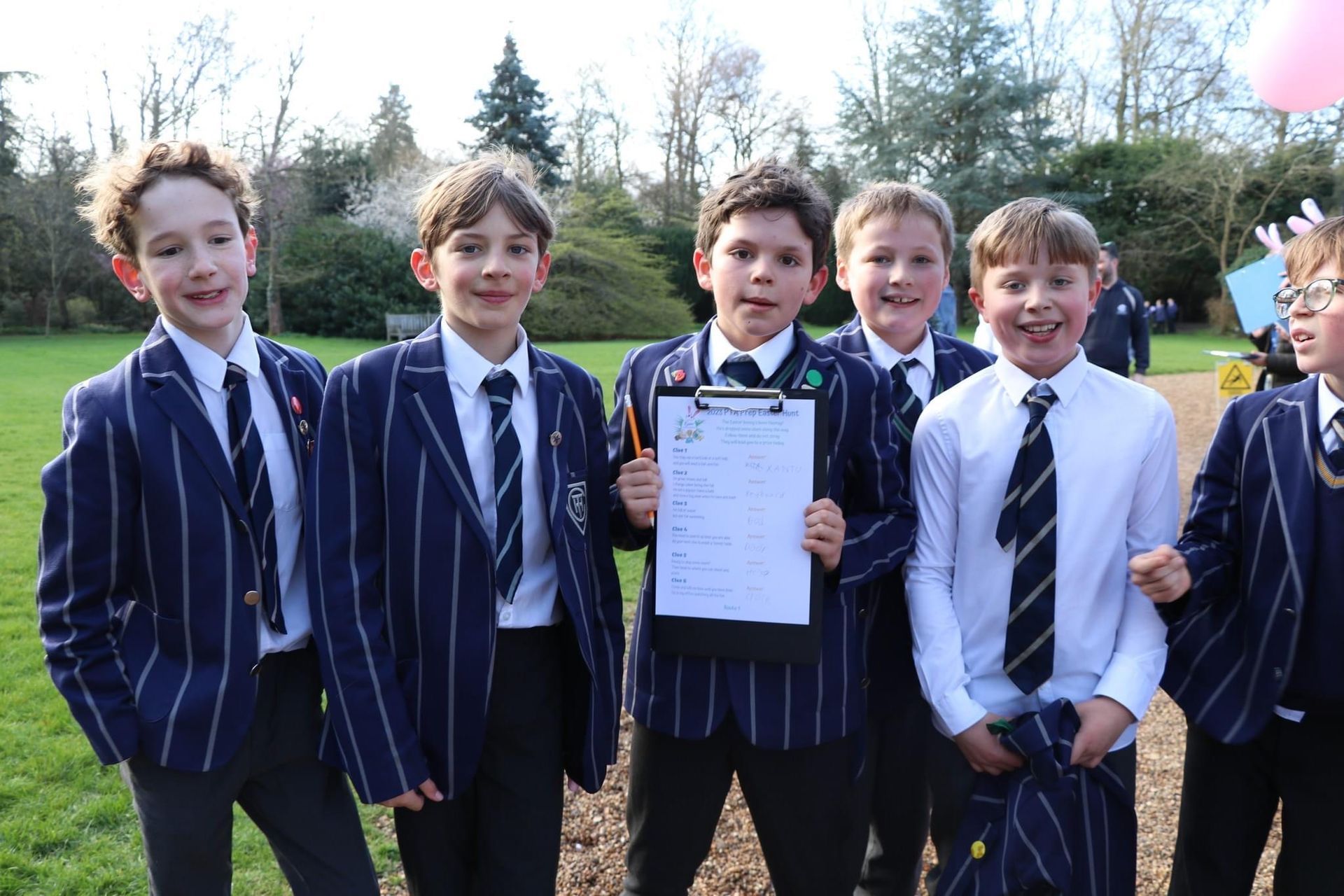 Boys in blazers holding clipboard, posing on a grassy field, smiling at the camera, trees in the background.