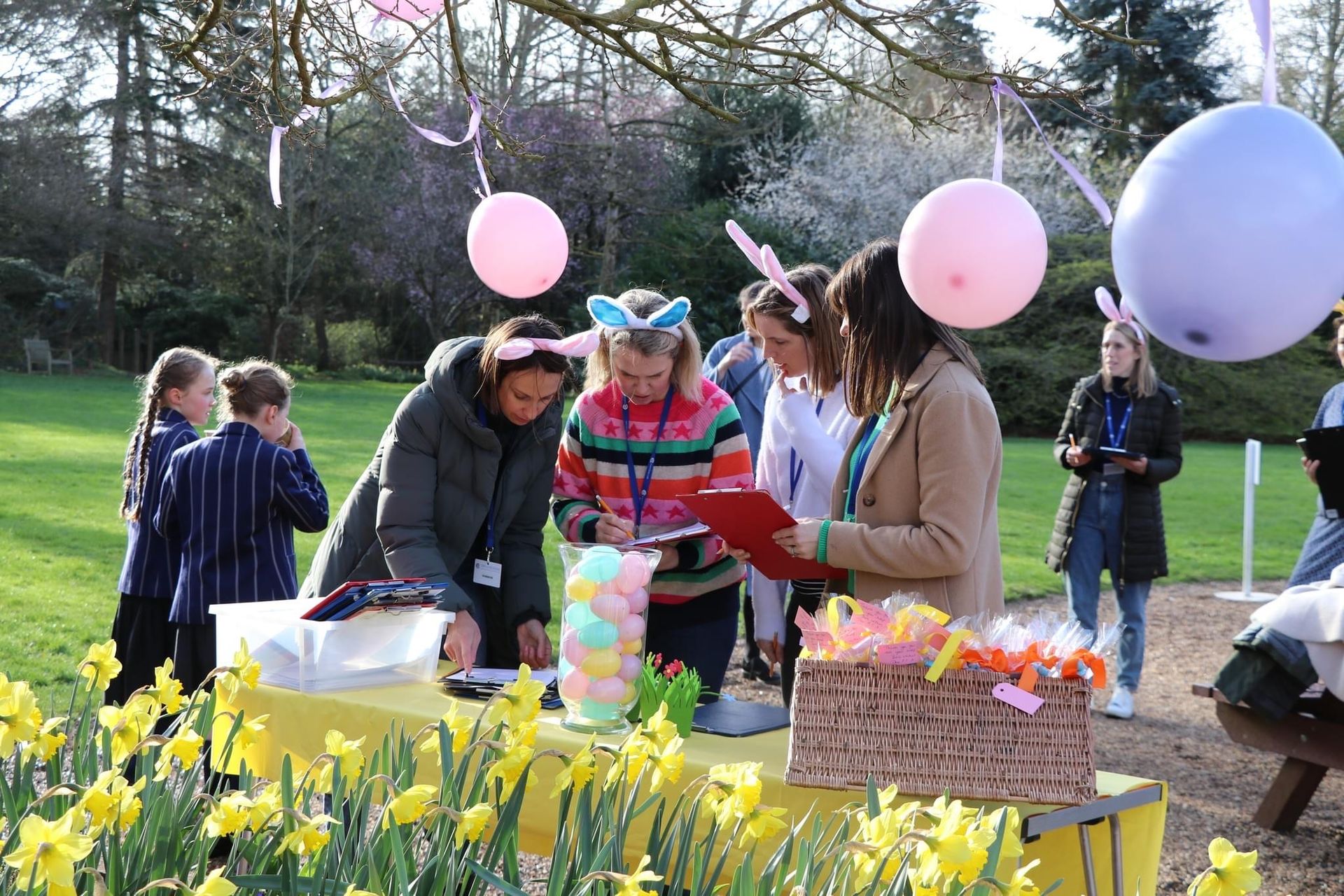 Easter egg hunt with children and adults, outdoor setting, balloons, daffodils, and participants wearing bunny ears.