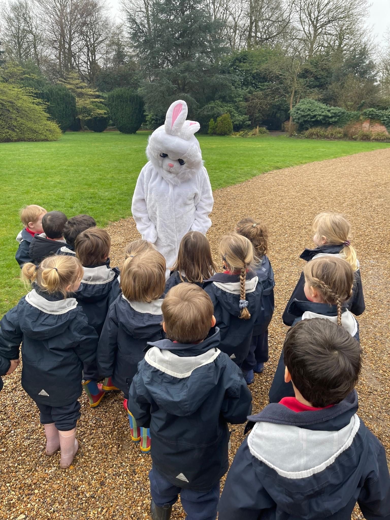 Children in raincoats gather to meet the Easter Bunny on a gravel path in a grassy park.