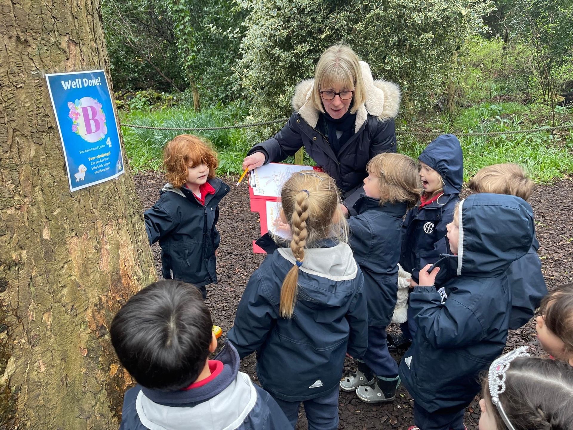 A teacher shows children something in a forest. Kids wear blue coats. A poster is on the tree.
