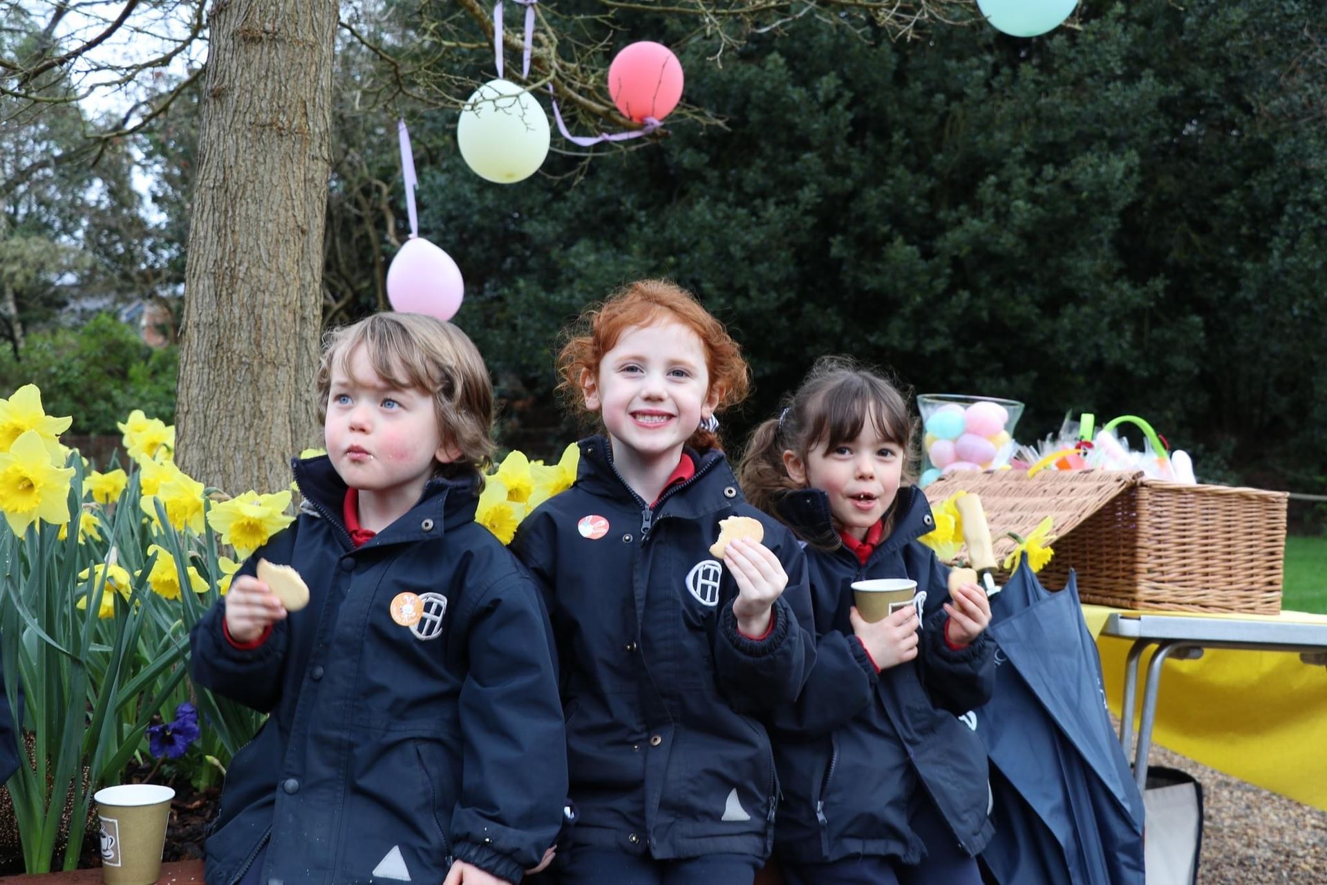 Three children in jackets eating cookies outdoors, smiling near daffodils and balloons.