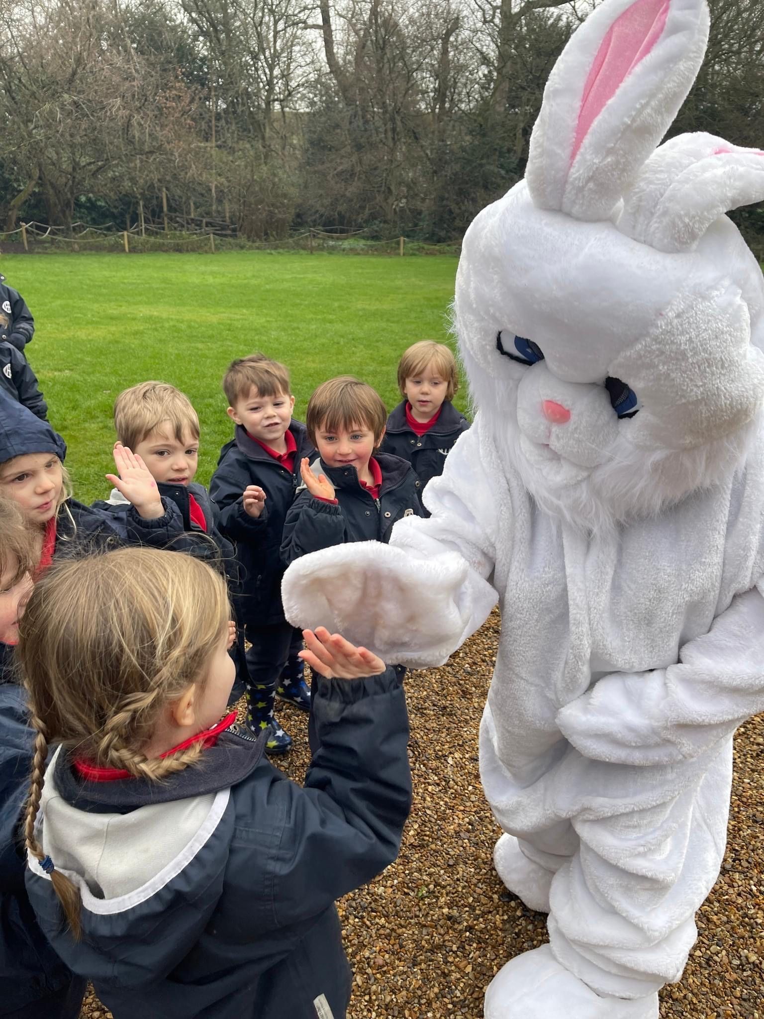 Children and Easter Bunny outdoors. Bunny gives a child something. Others clap. Green grass, trees.
