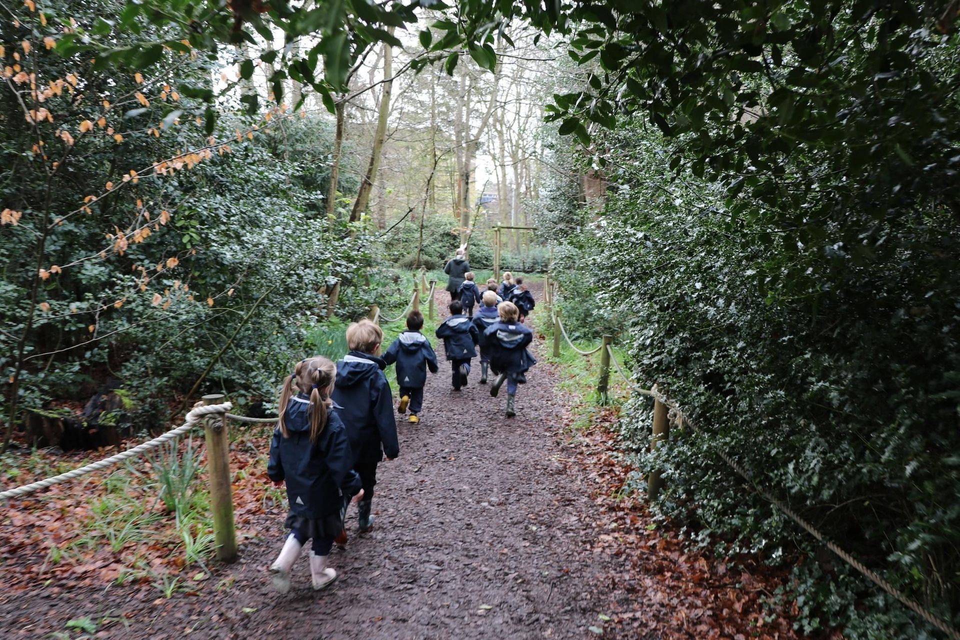 Children in uniform walking on a muddy forest path, surrounded by green bushes and trees.