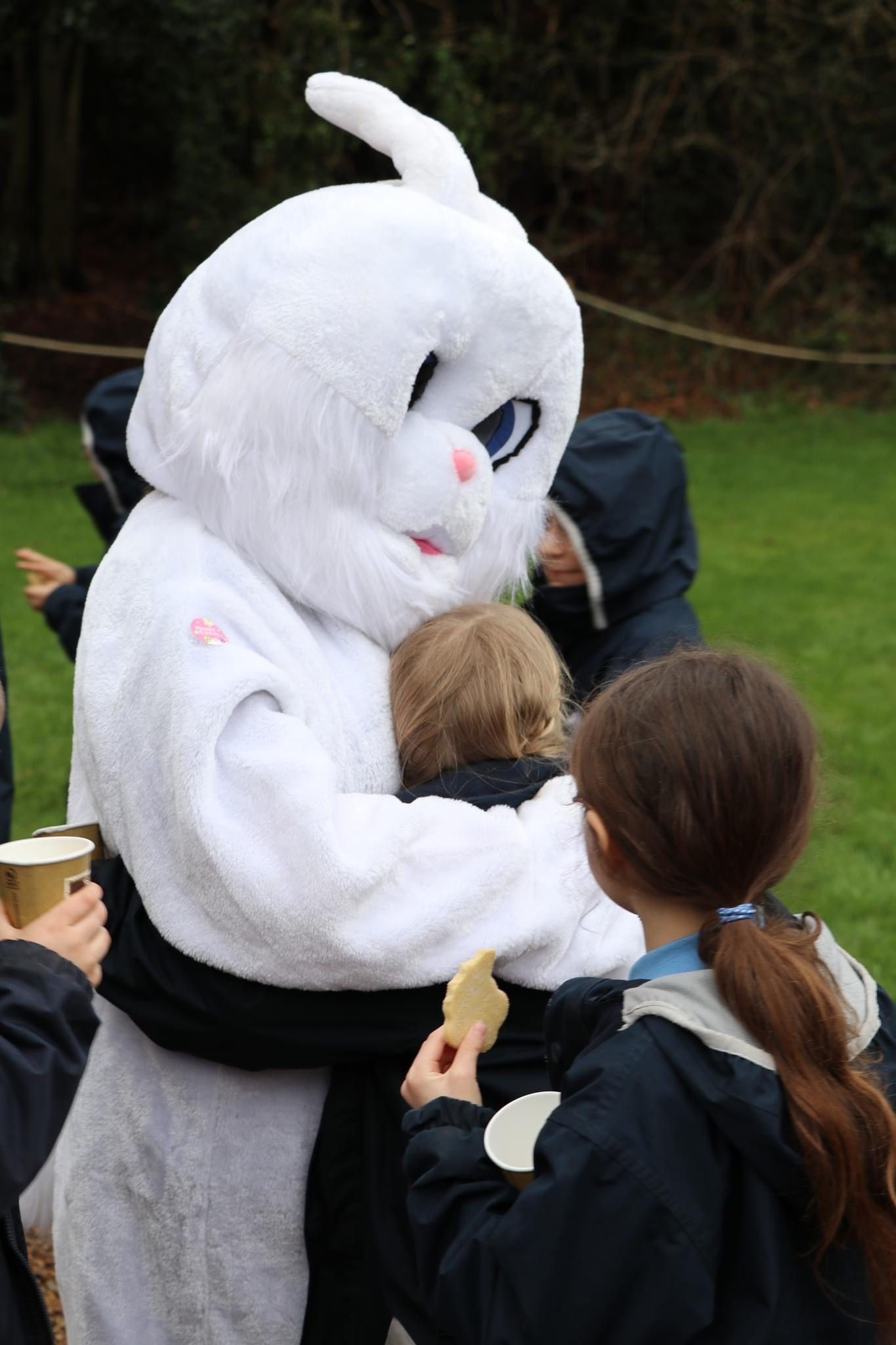 White bunny mascot hugging a child; children in the background. Outdoor setting.