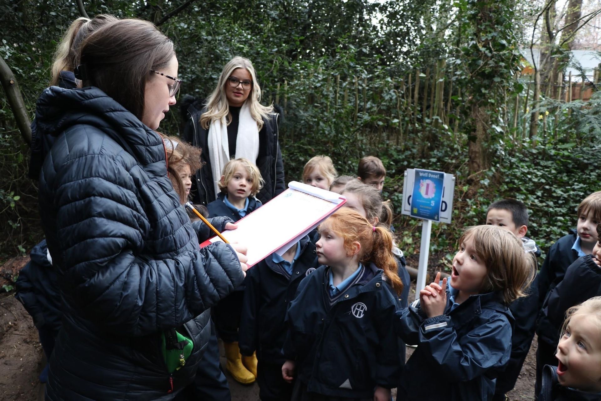 Teacher with clipboard instructing children in outdoor setting; another woman watches nearby.