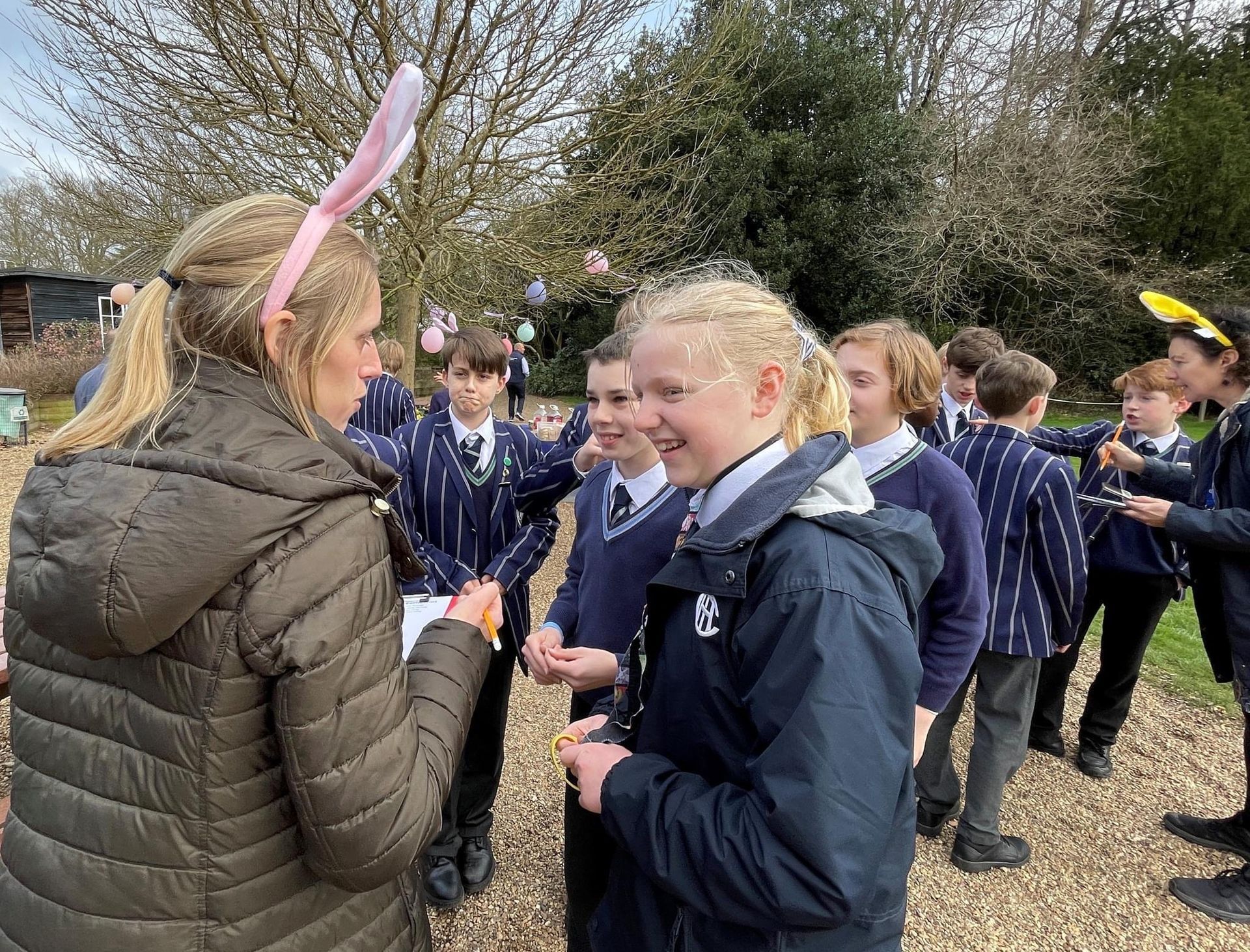 Woman in bunny ears talks with student; group of students outdoors in school uniforms.