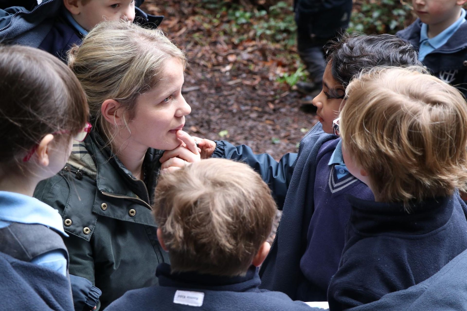 Teacher in a forest talks with a group of school children, blue uniforms and jackets, near trees.