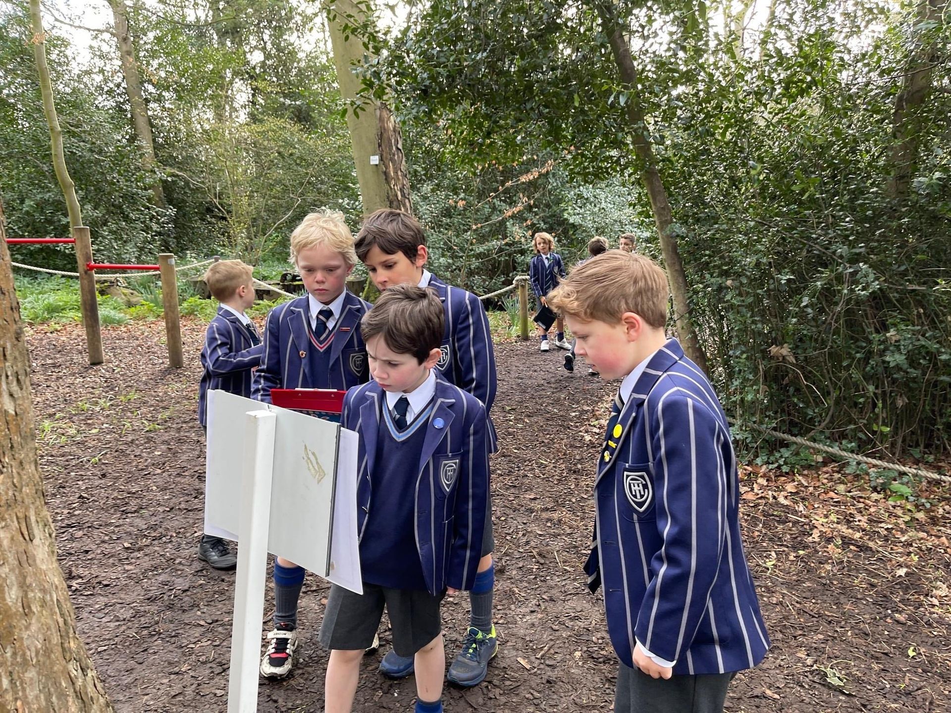 Boys in school uniforms reading signs on a muddy trail in a wooded area.