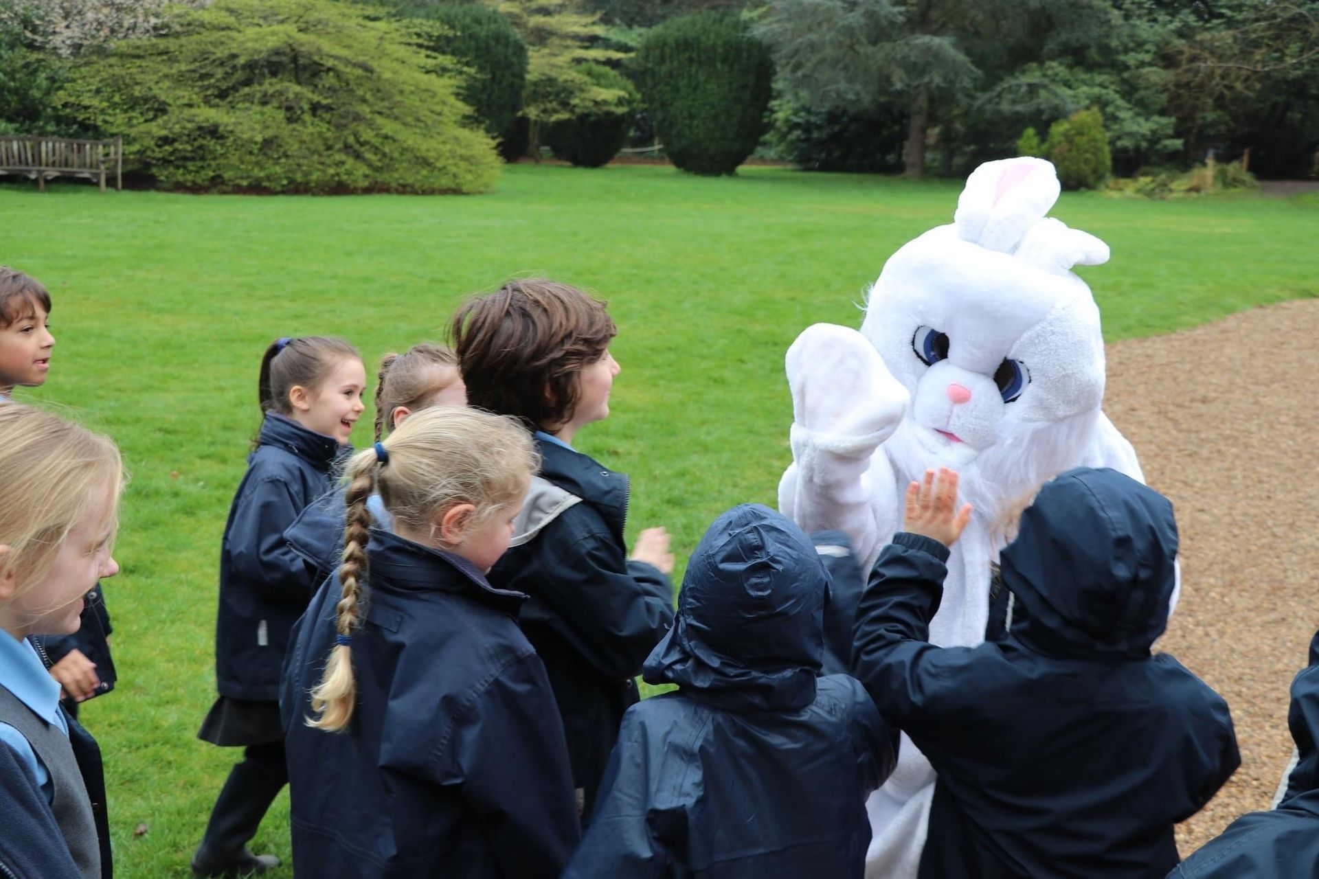 Children in school uniforms greet a white Easter Bunny costume in a park setting.