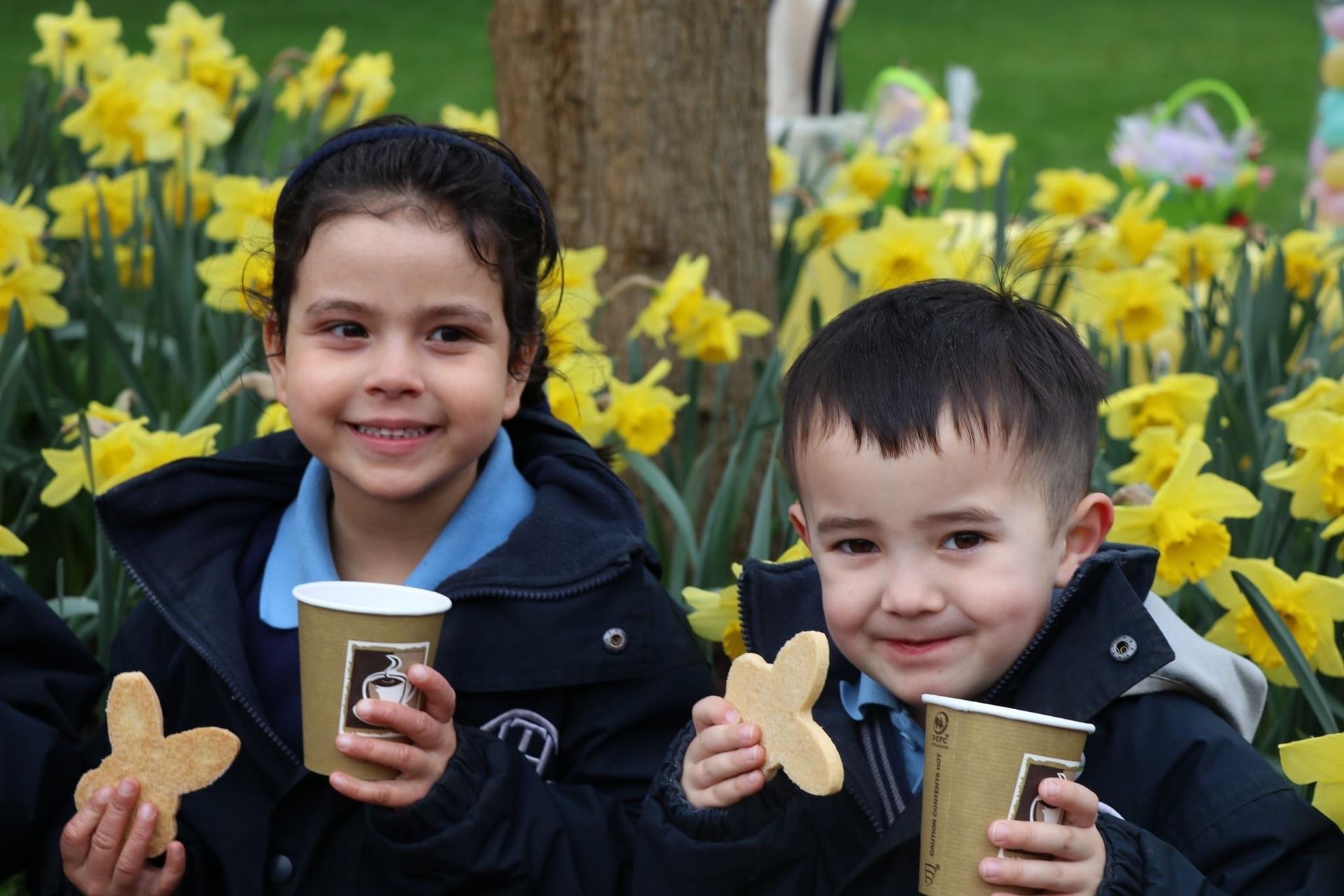 Two children in school uniforms holding cookies and drinks in front of yellow daffodils.