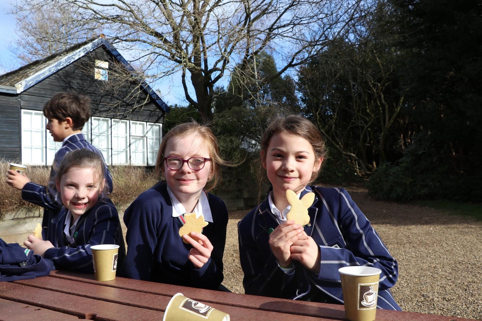 Children in school uniforms, holding bunny cookies, smile at a table outdoors. A building and trees are in the background.