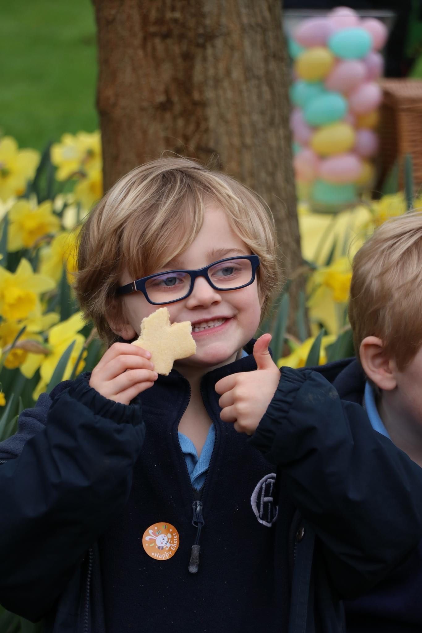 Boy with glasses, eating a cookie and giving a thumbs up. Daffodils and balloons in background.