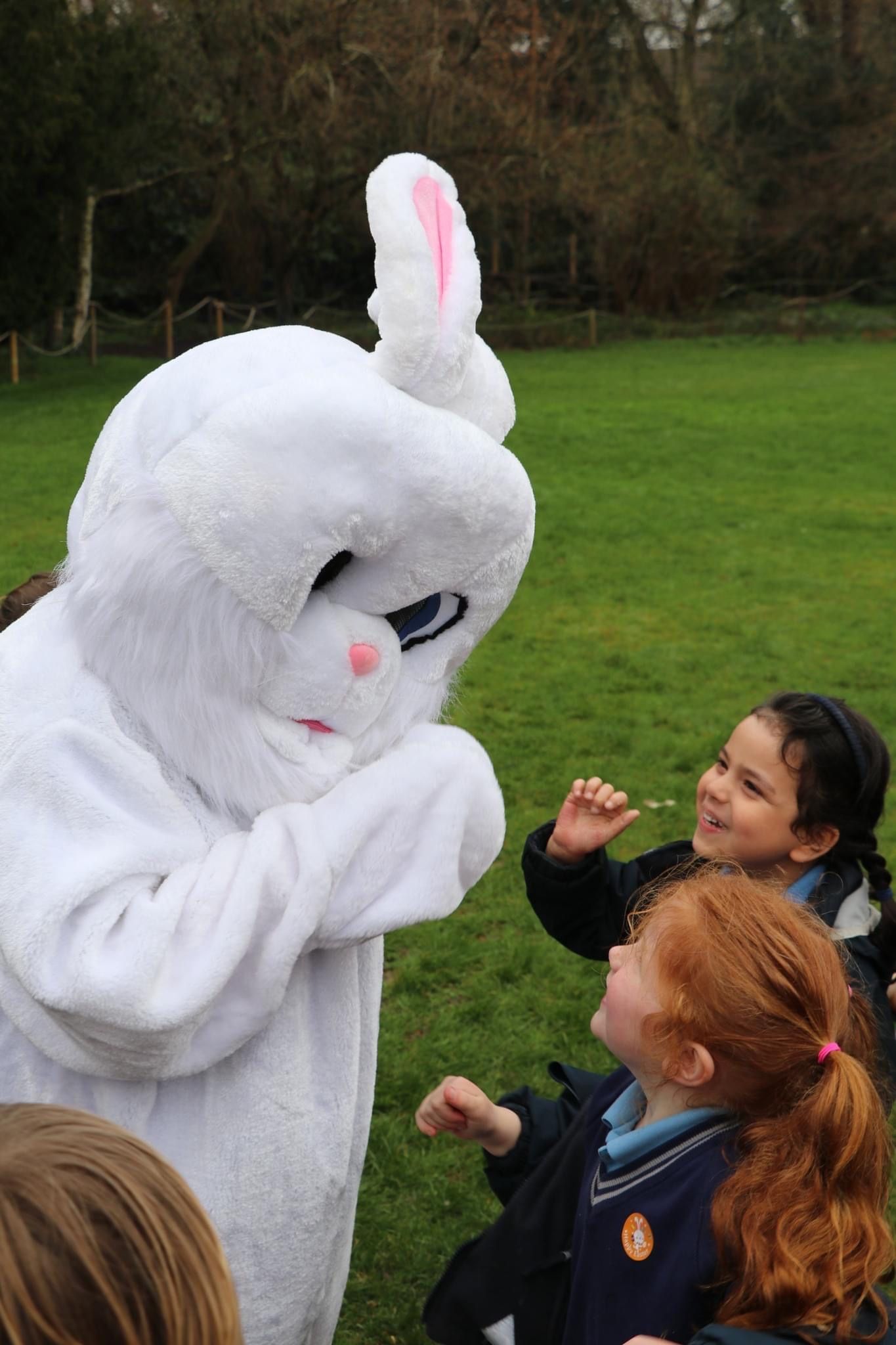 A person in a white Easter bunny costume interacting with two children outdoors in a park.