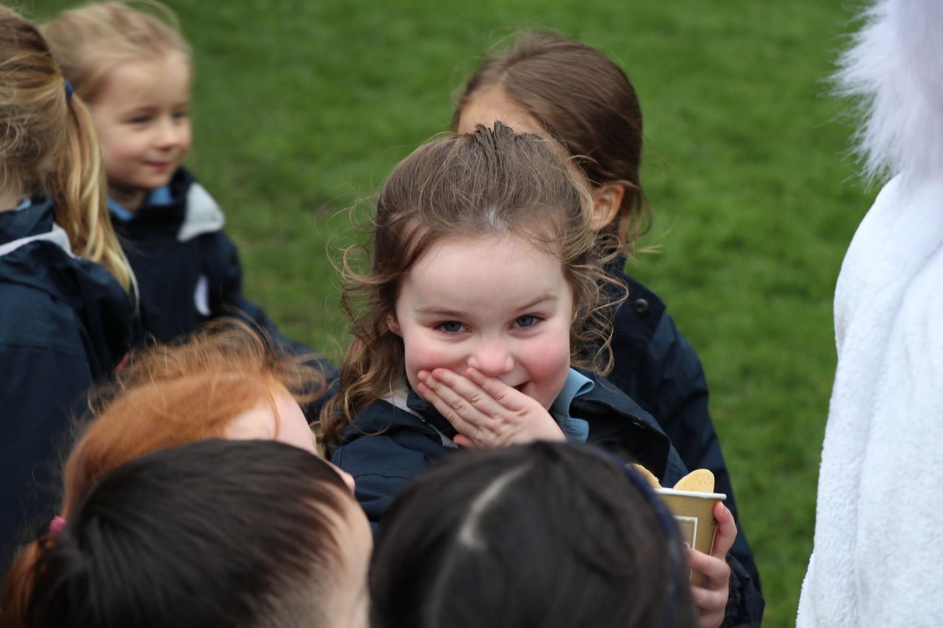 Girl with a hand over her mouth laughs, surrounded by other children wearing school uniforms outdoors.