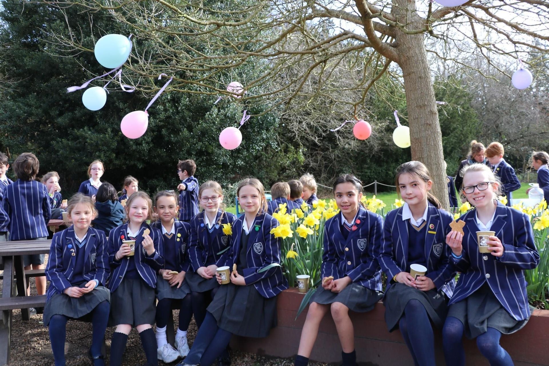 Group of schoolgirls outside, some holding treats, tree with balloons, daffodils.