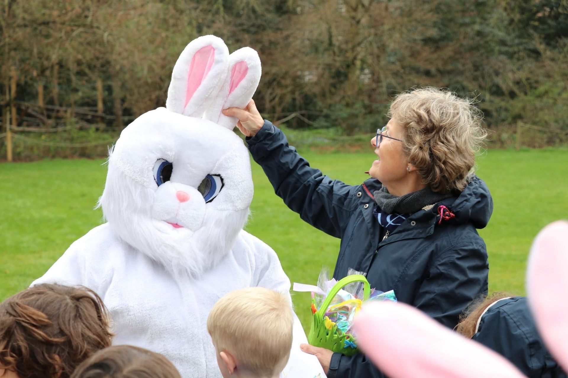 Woman adjusts Easter Bunny's ear outside. Children with Easter baskets watch. Green grass, trees in background.