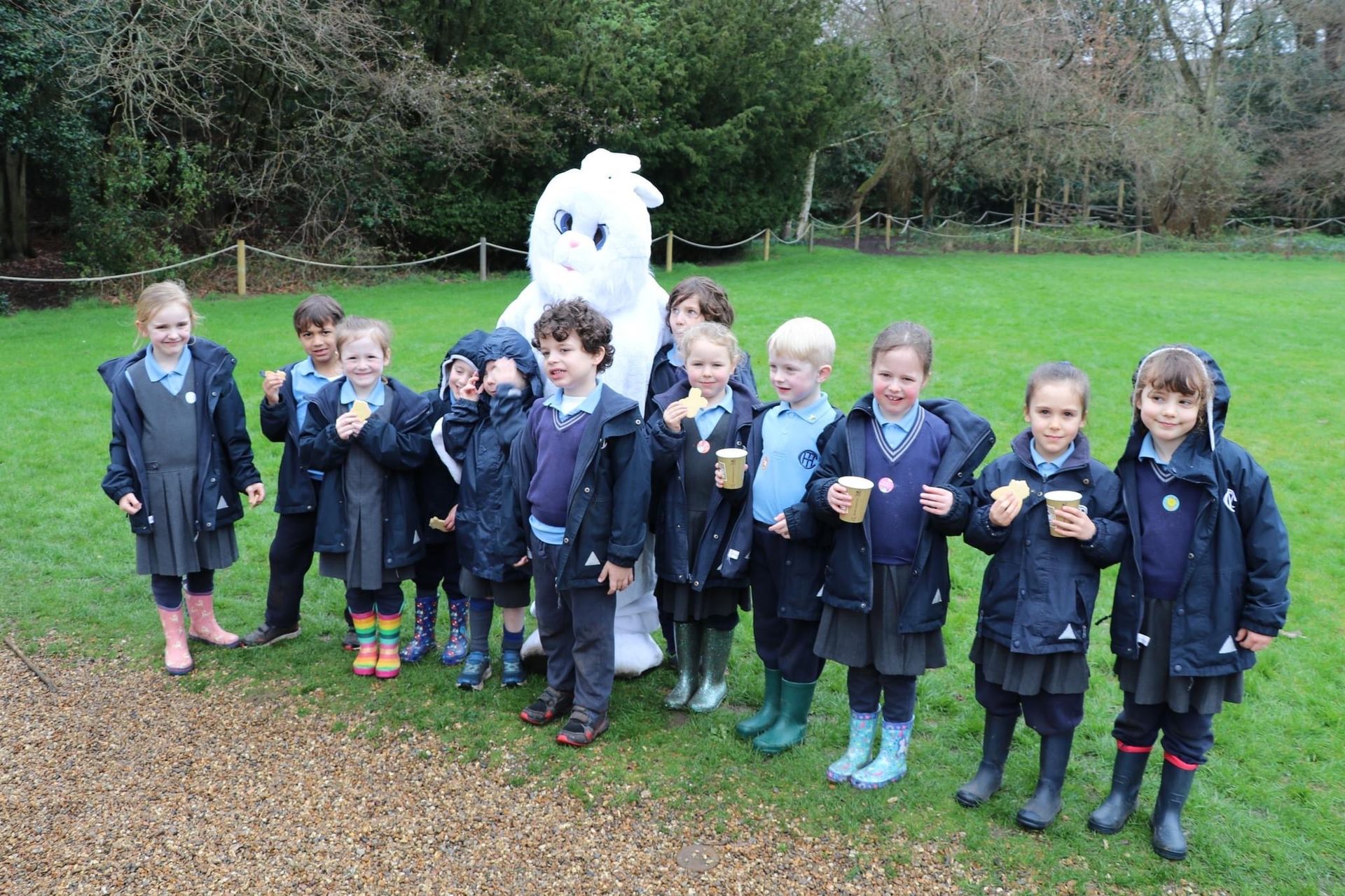 Children in school uniforms with a mascot outdoors, all holding drinks.