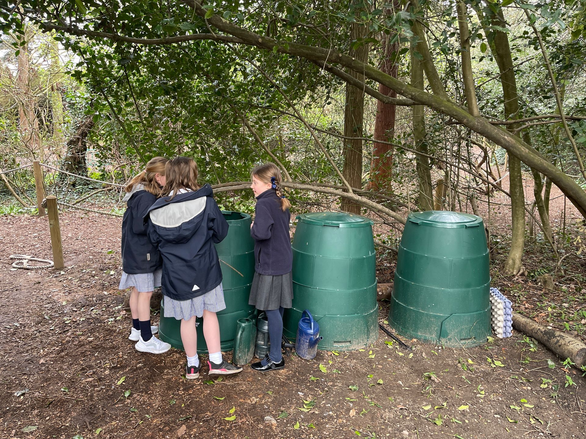 Children by green compost bins in a wooded area.