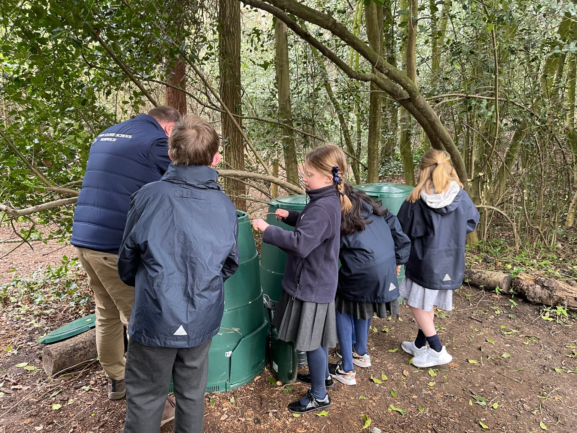 Children and an adult examine a green bin in a wooded area.