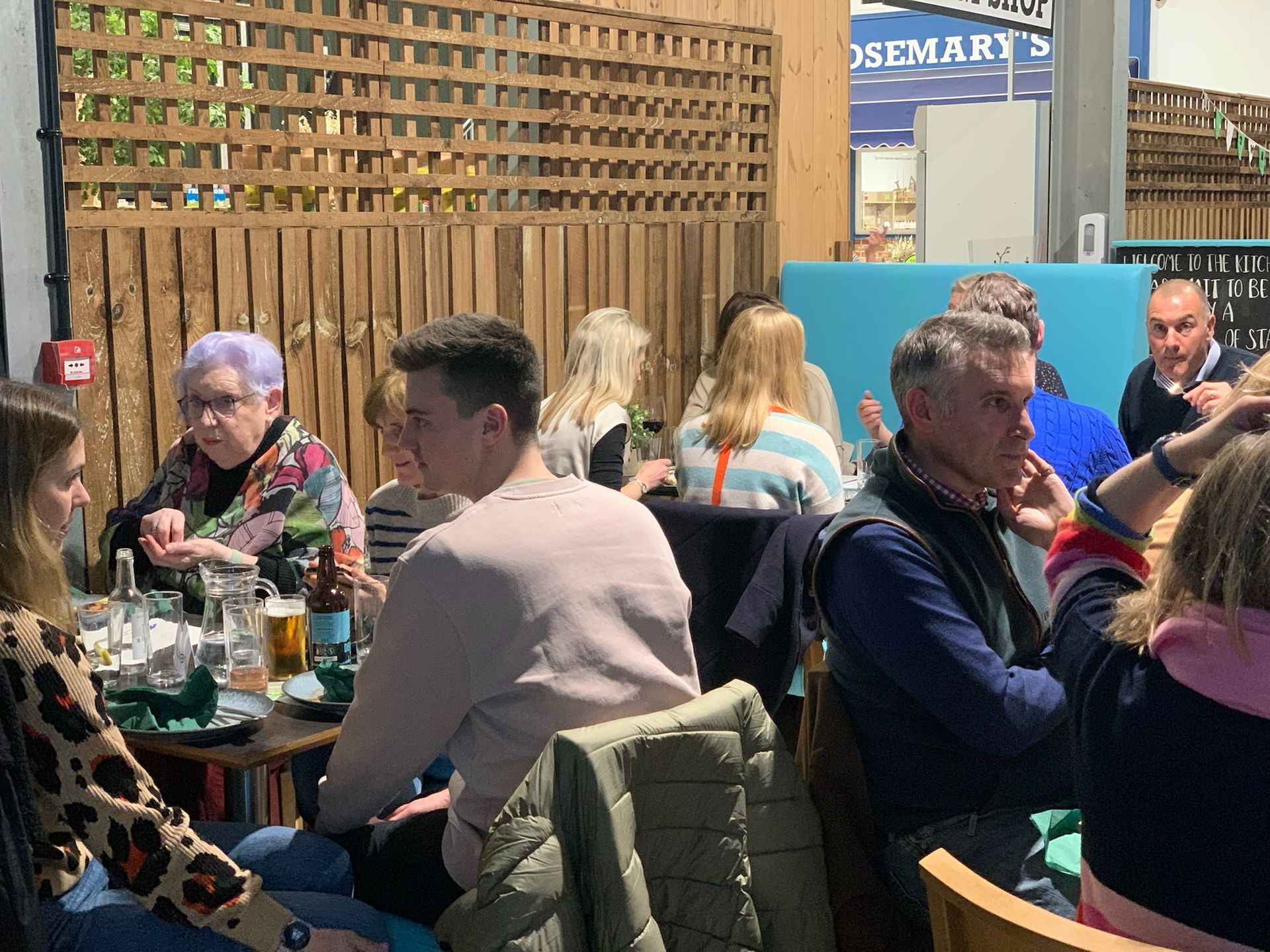 People seated at tables in a cafe; some drinking, socializing. Wooden paneling and blue walls visible.