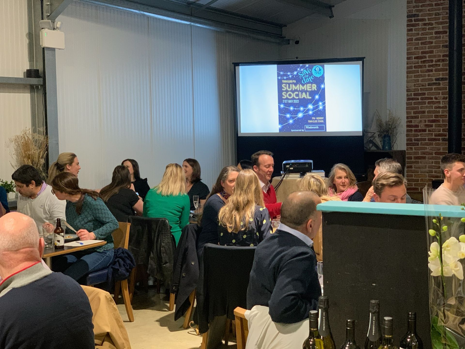 People at an event, seated around tables, watching a screen with a 