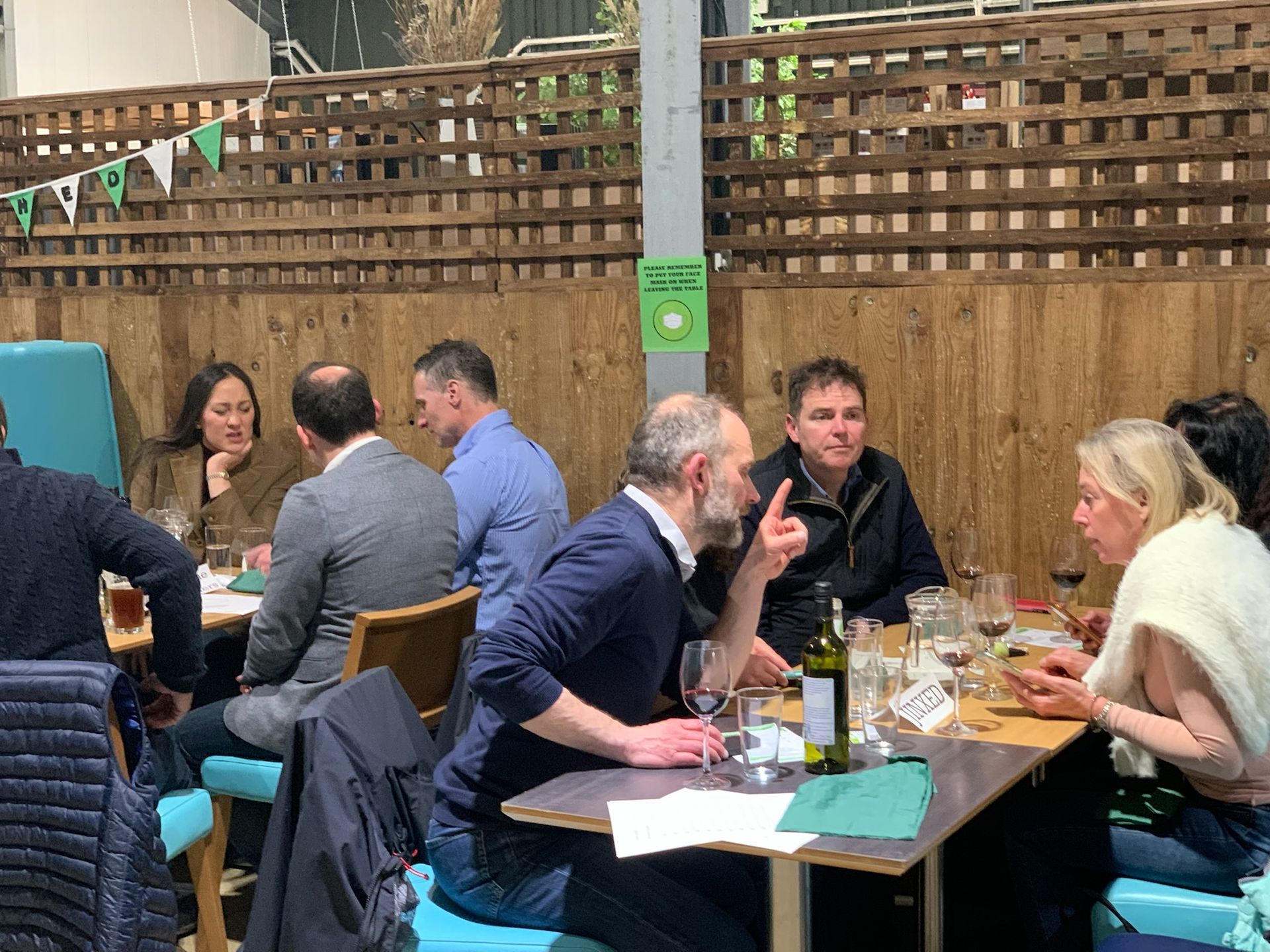 Group of people at a table, drinking and talking outdoors. Wooden fence and teal seating in the background.