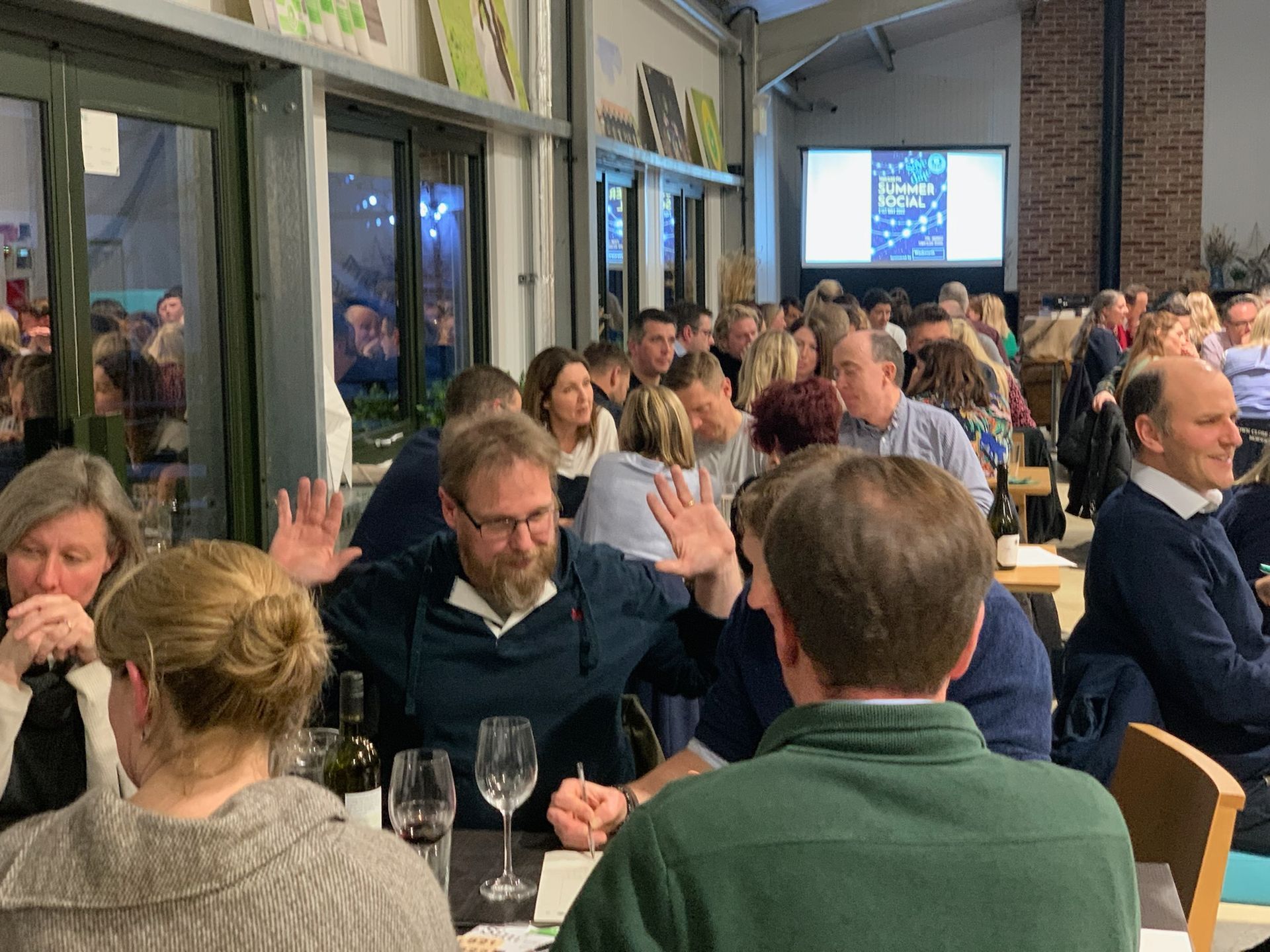 People at tables in a restaurant, one man gesturing with hands, others drinking wine.