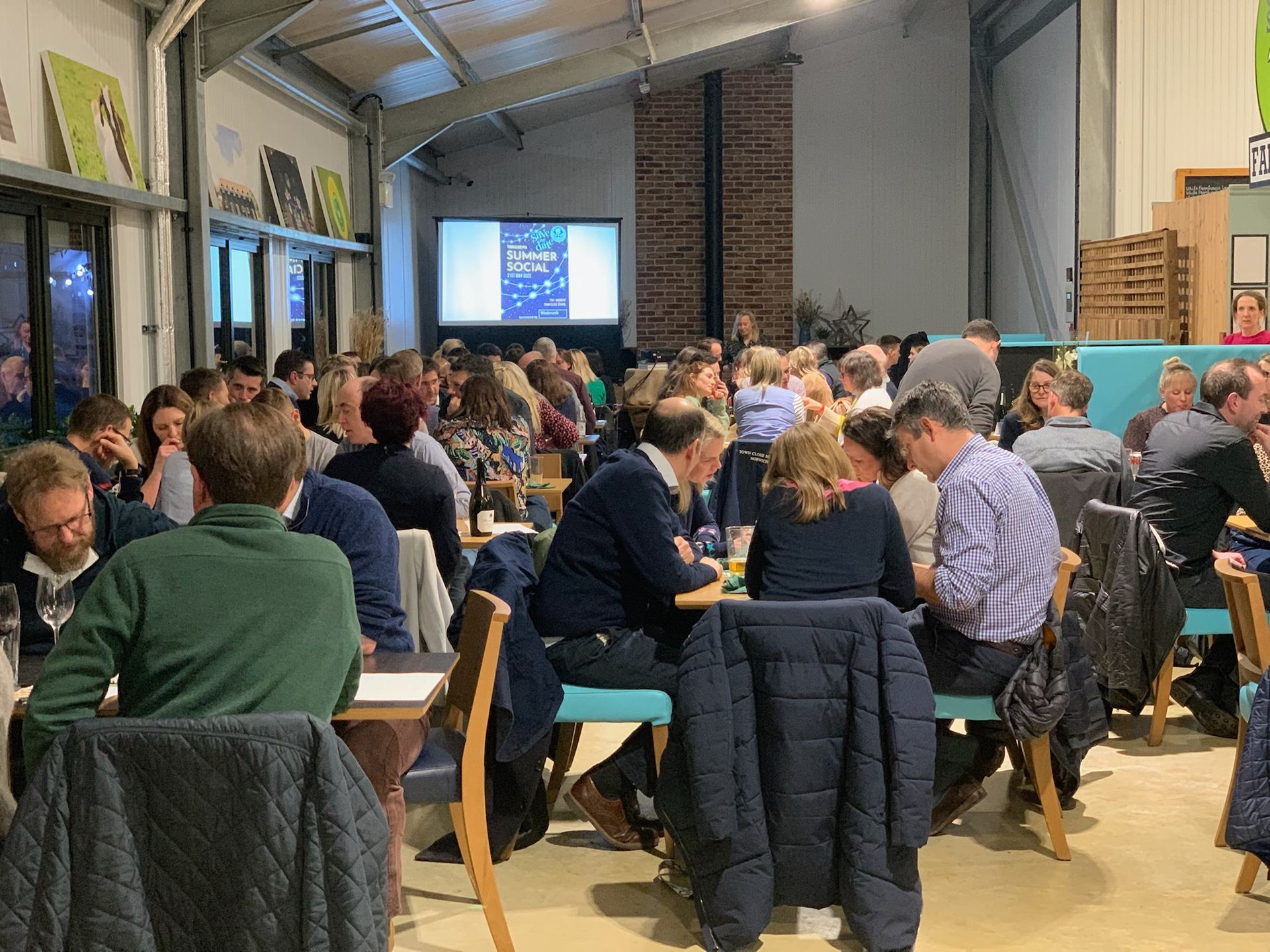 Large group of people seated at tables in a hall, interacting and listening; a screen displays text.