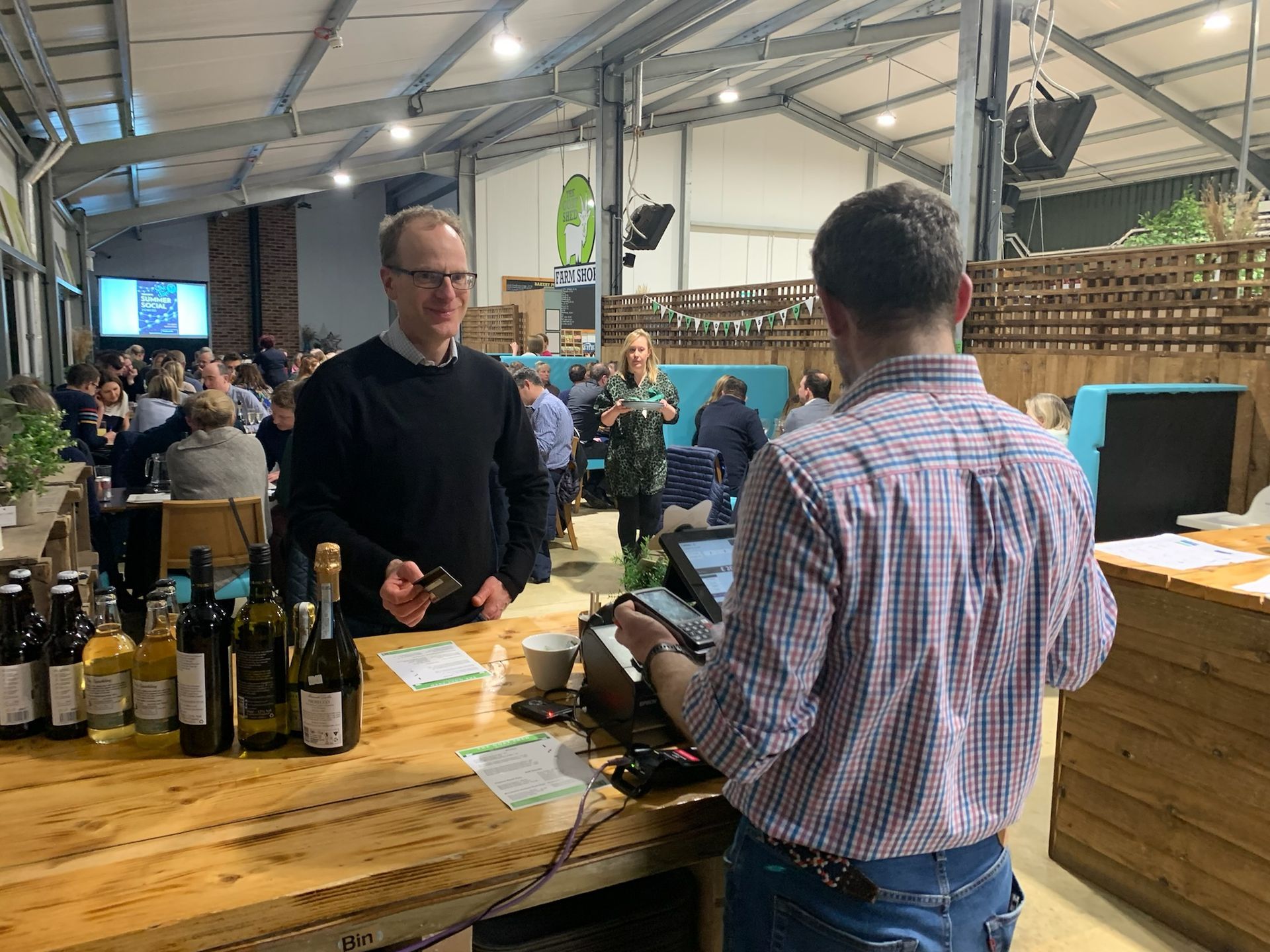 Man buying wine at a tasting event. Person behind the counter, other people at tables in the background.