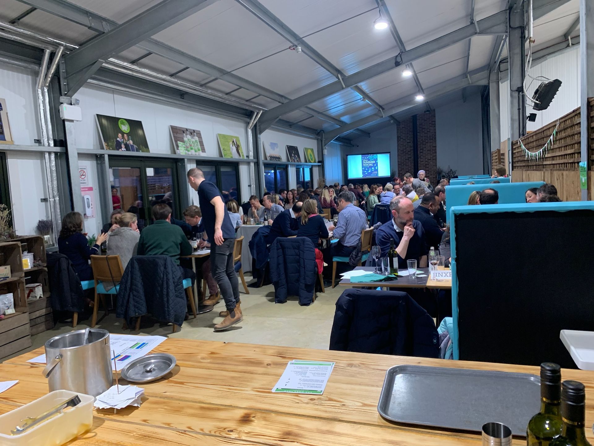Indoor event: People seated at tables, eating and interacting. Servers, wood bar, overhead lighting.