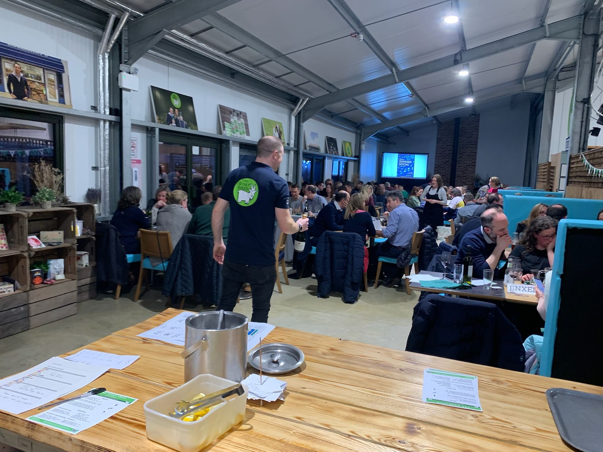 Restaurant scene: A man in a black shirt addresses a crowd seated at tables. Indoor setting with art on the walls.